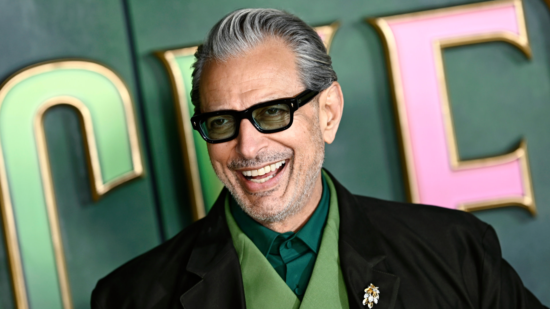 Jeff Goldblum, wearing glasses and a green outfit, smiles at an event with a colorful background.