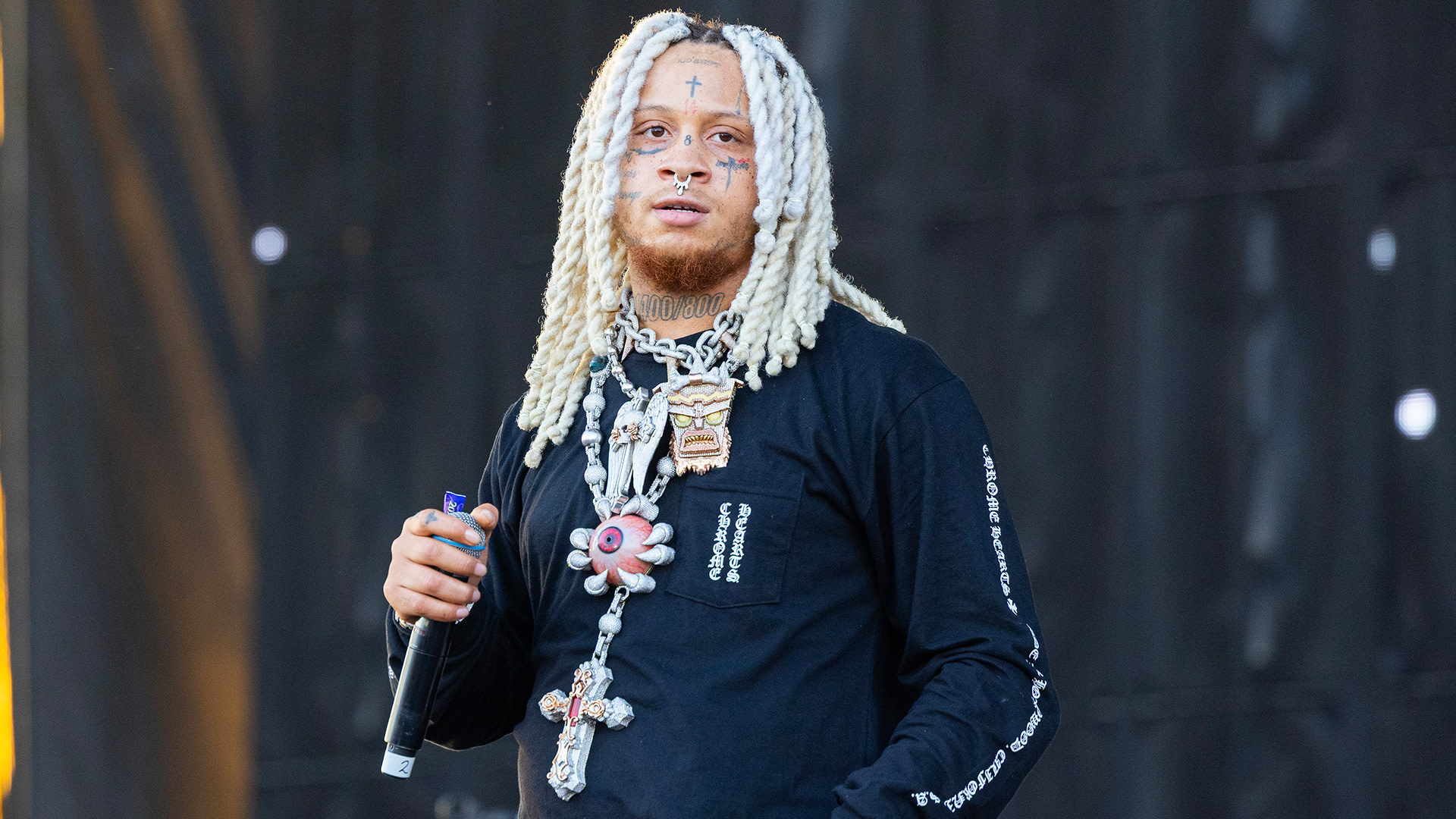 Rapper with long white dreadlocks and facial tattoos, holding a microphone on stage, wearing a black shirt and large necklaces.