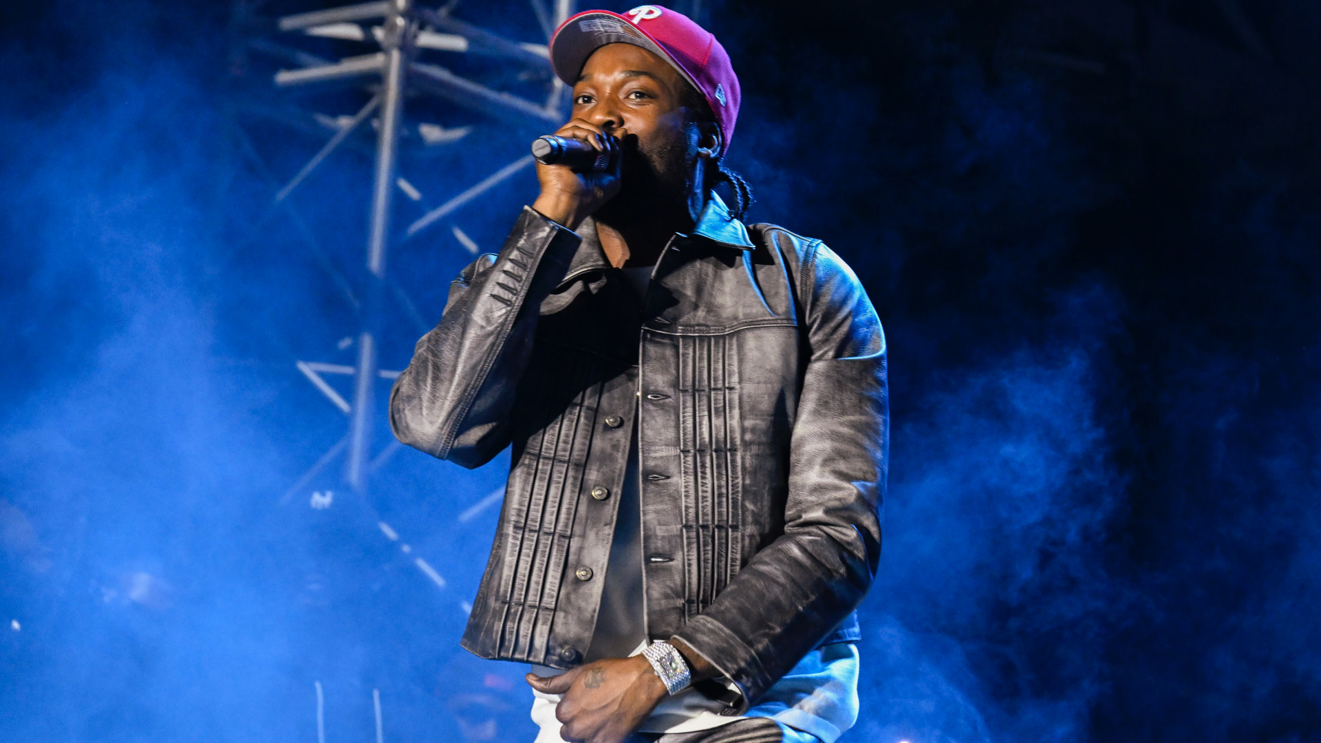 Meek Mill in a red cap and leather jacket performs on stage, holding a microphone, with blue smoke in the background.