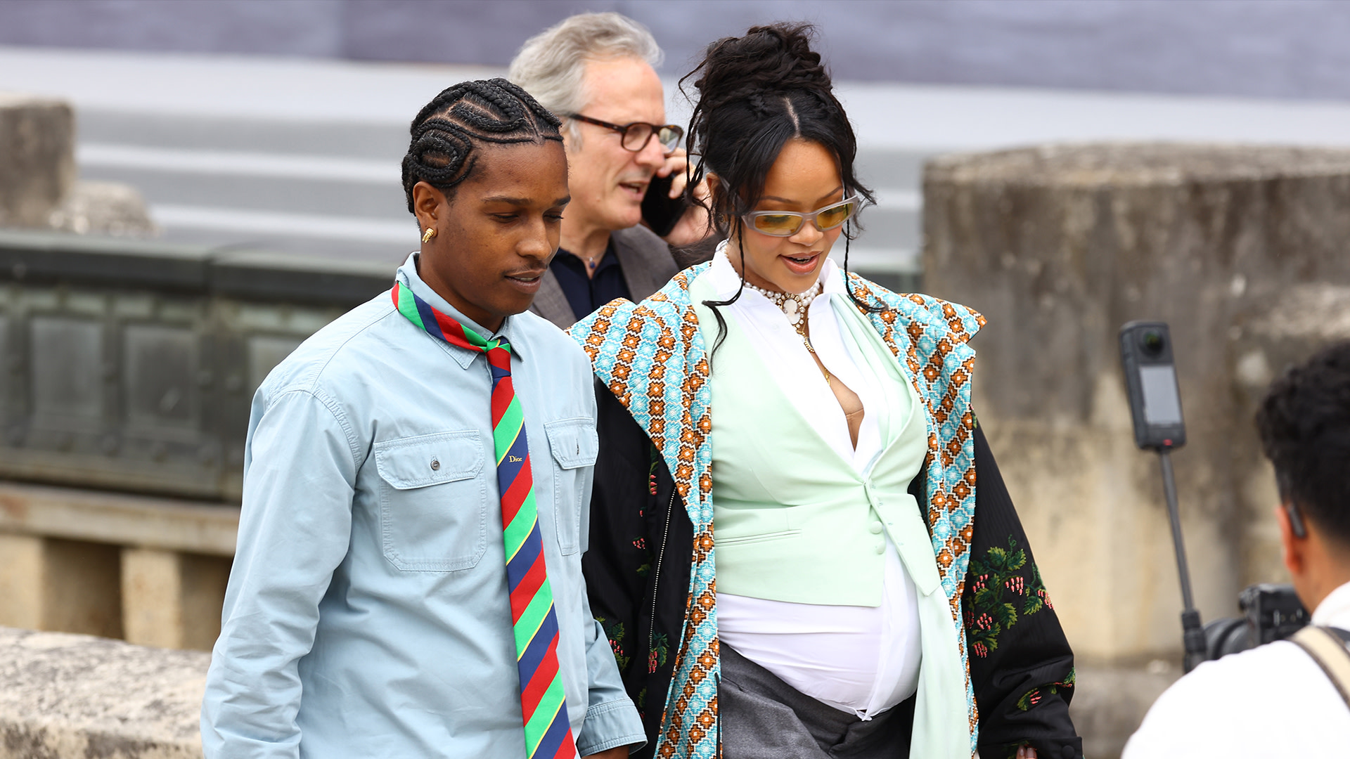 A$AP Rocky and Rihanna walking together. A$AP Rocky wears a blue shirt and striped tie, Rihanna wears a patterned coat and sunglasses.