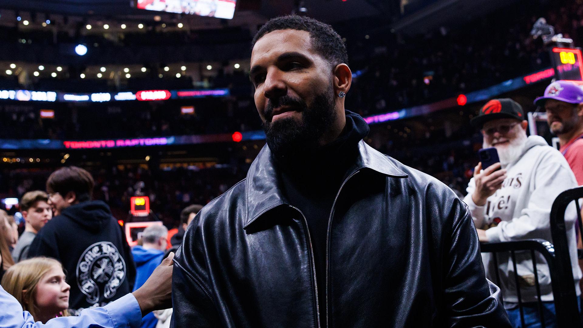 Drake in a black leather jacket at a Toronto Raptors game, surrounded by fans in an arena.