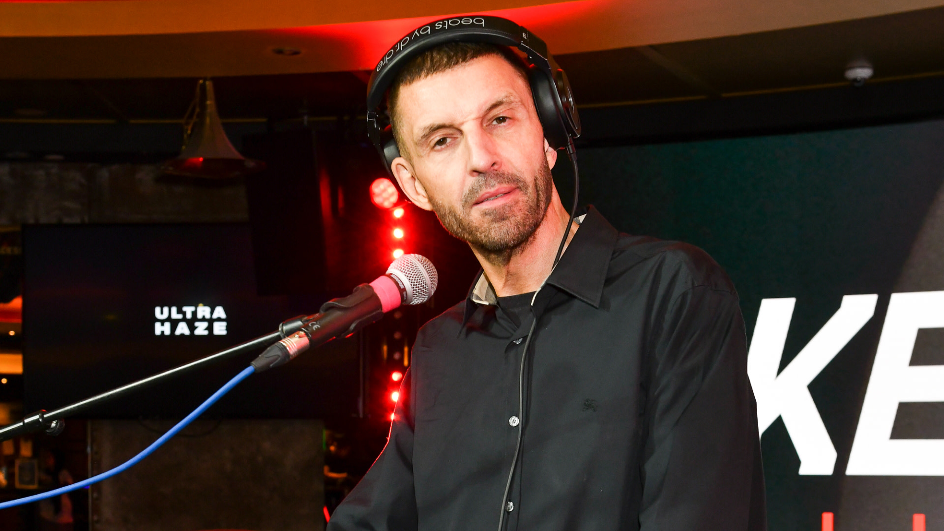 A DJ wearing headphones stands at a microphone, with a background displaying "Ultra Haze." The setting appears to be a music event.