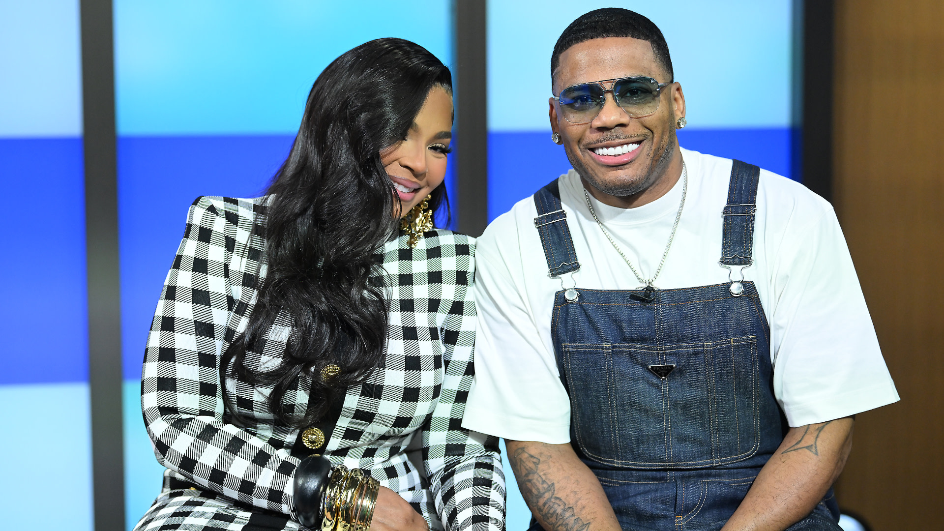 Ashanti in a black and white checkered outfit and Nelly in denim overalls smiling together.