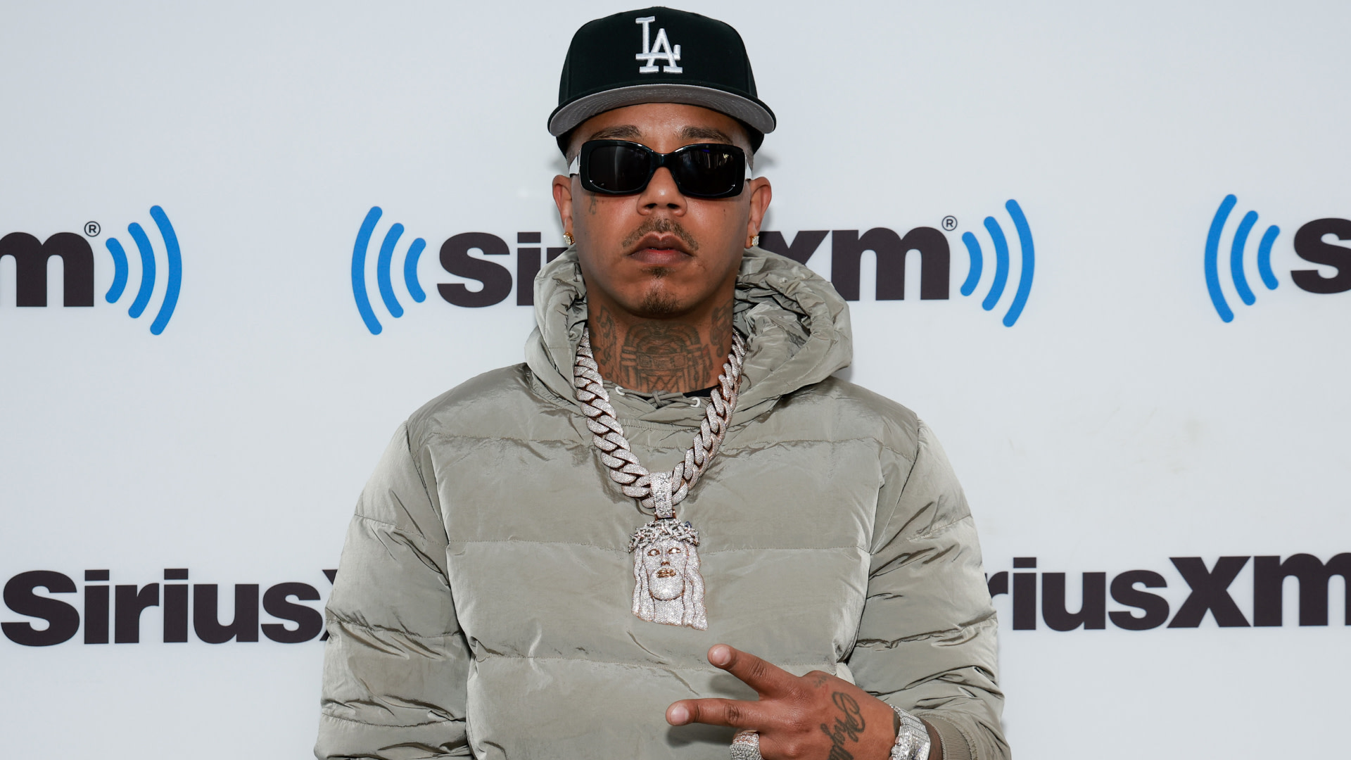 Hitmaka in sunglasses, a black LA cap, a gray puffer jacket, and a large chain necklace, posing at a SiriusXM event.