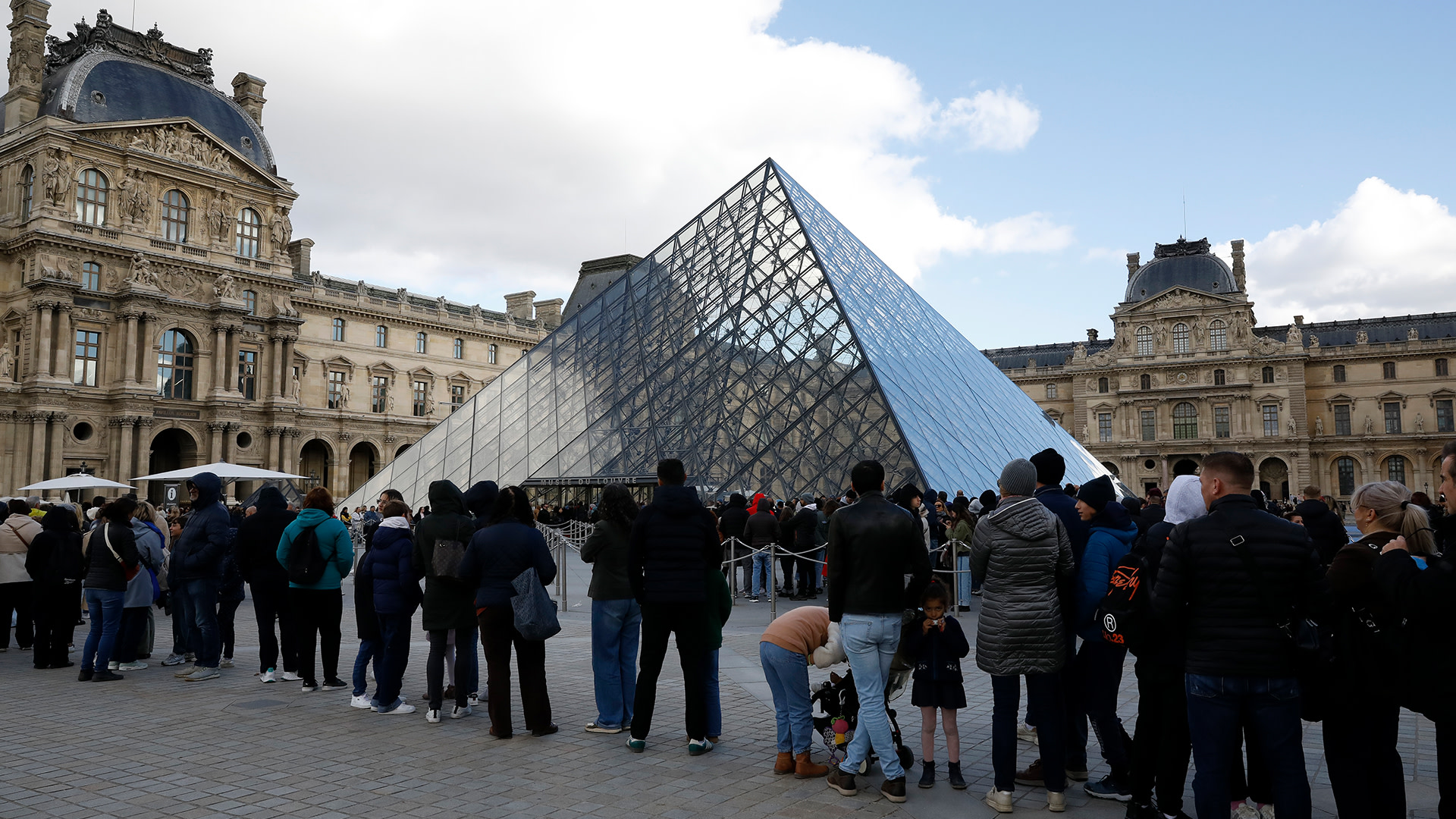 People in line outside the Louvre Pyramid in Paris, with the historic museum buildings in the background under a partly cloudy sky.
