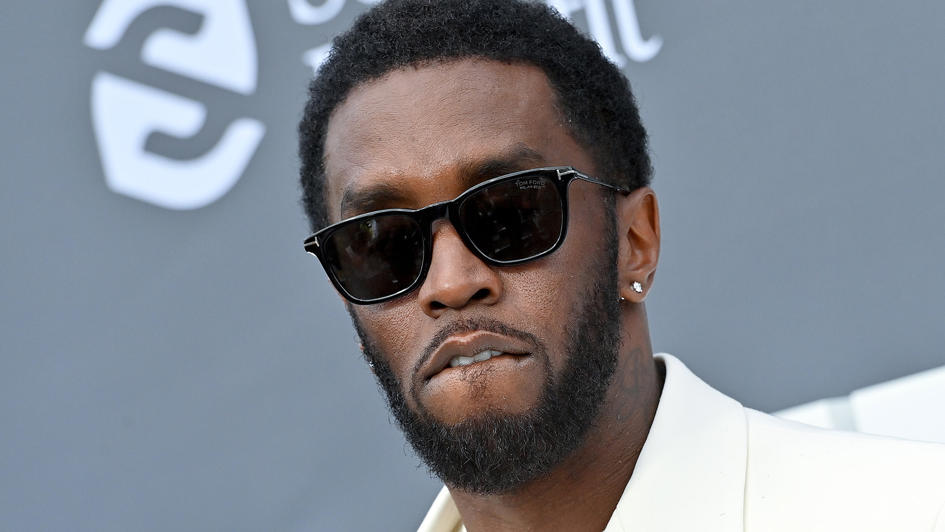 Sean "Diddy" Combs wearing sunglasses, with a trimmed beard, in a white suit, biting his lip.