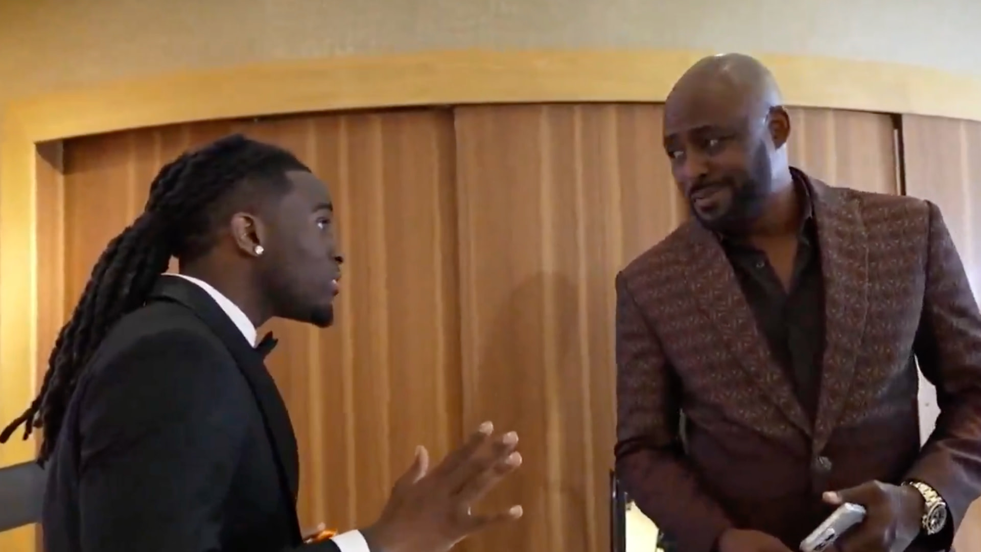 Two men in formal attire are engaged in conversation. One has long hair, and the other is bald with a beard, holding a phone.