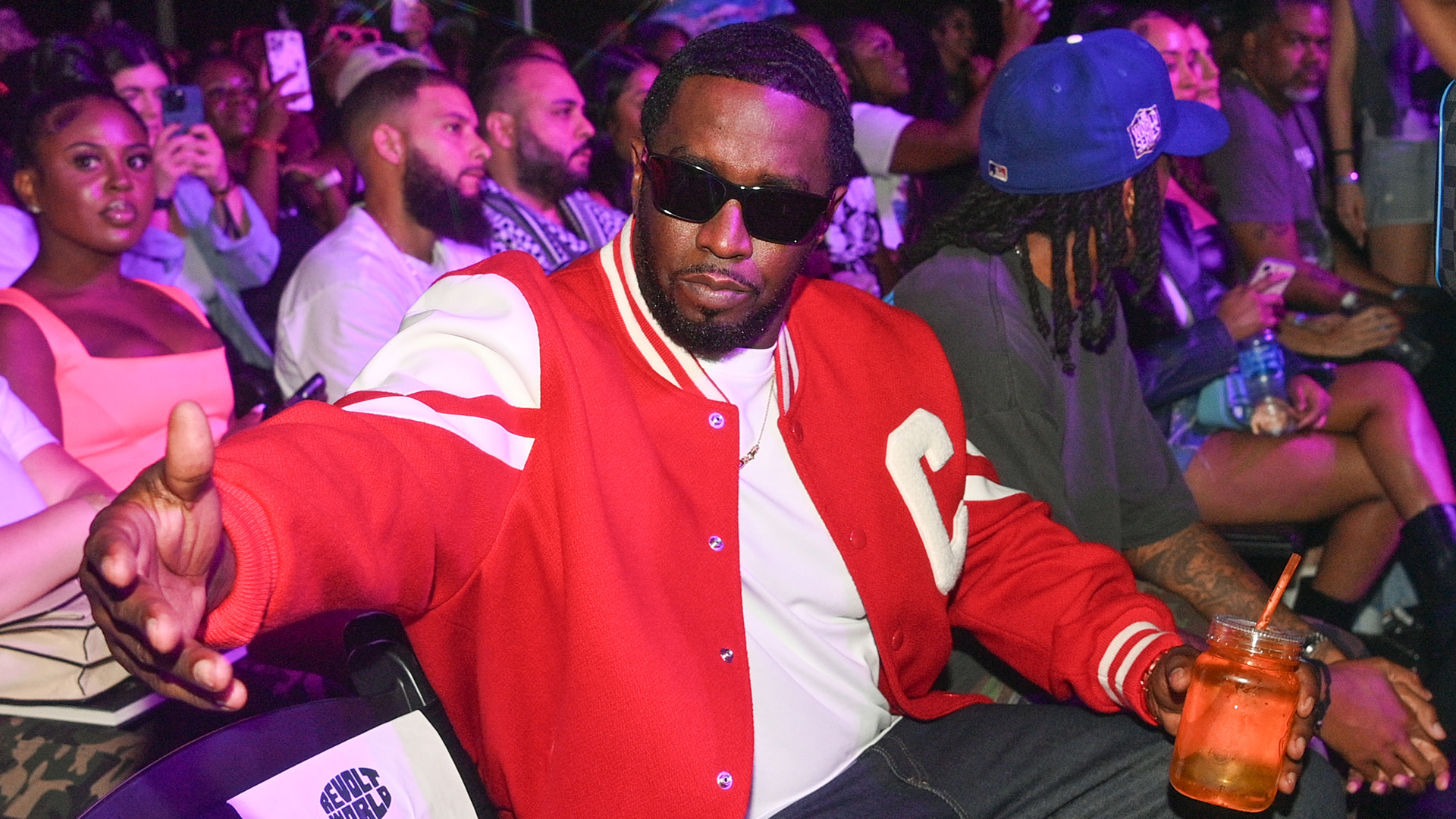 Sean "Diddy" Combs wearing sunglasses and a red jacket, sitting in a lively crowd at an event.