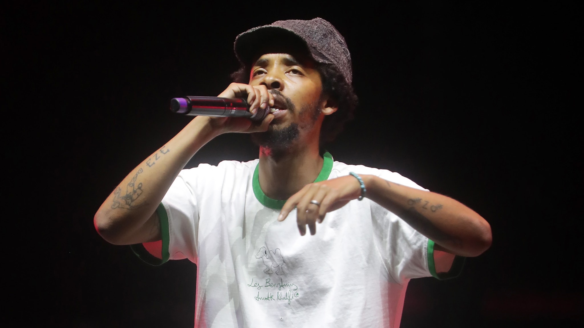 Earl Sweatshirt wearing a white t-shirt and a gray cap is performing on stage with a microphone in hand.