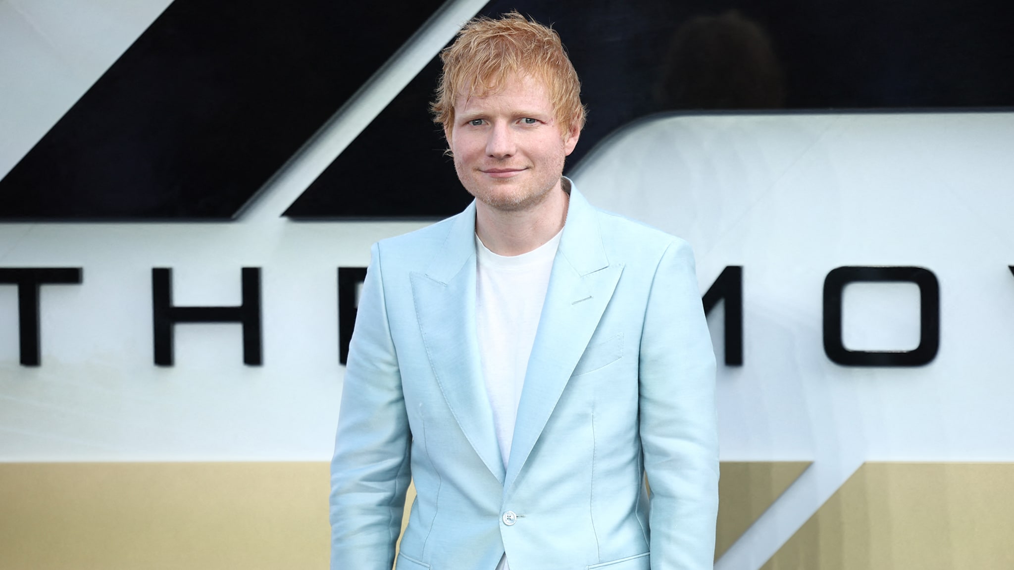 Ed Sheeran in a light blue suit, standing in front of a sign with bold black text.