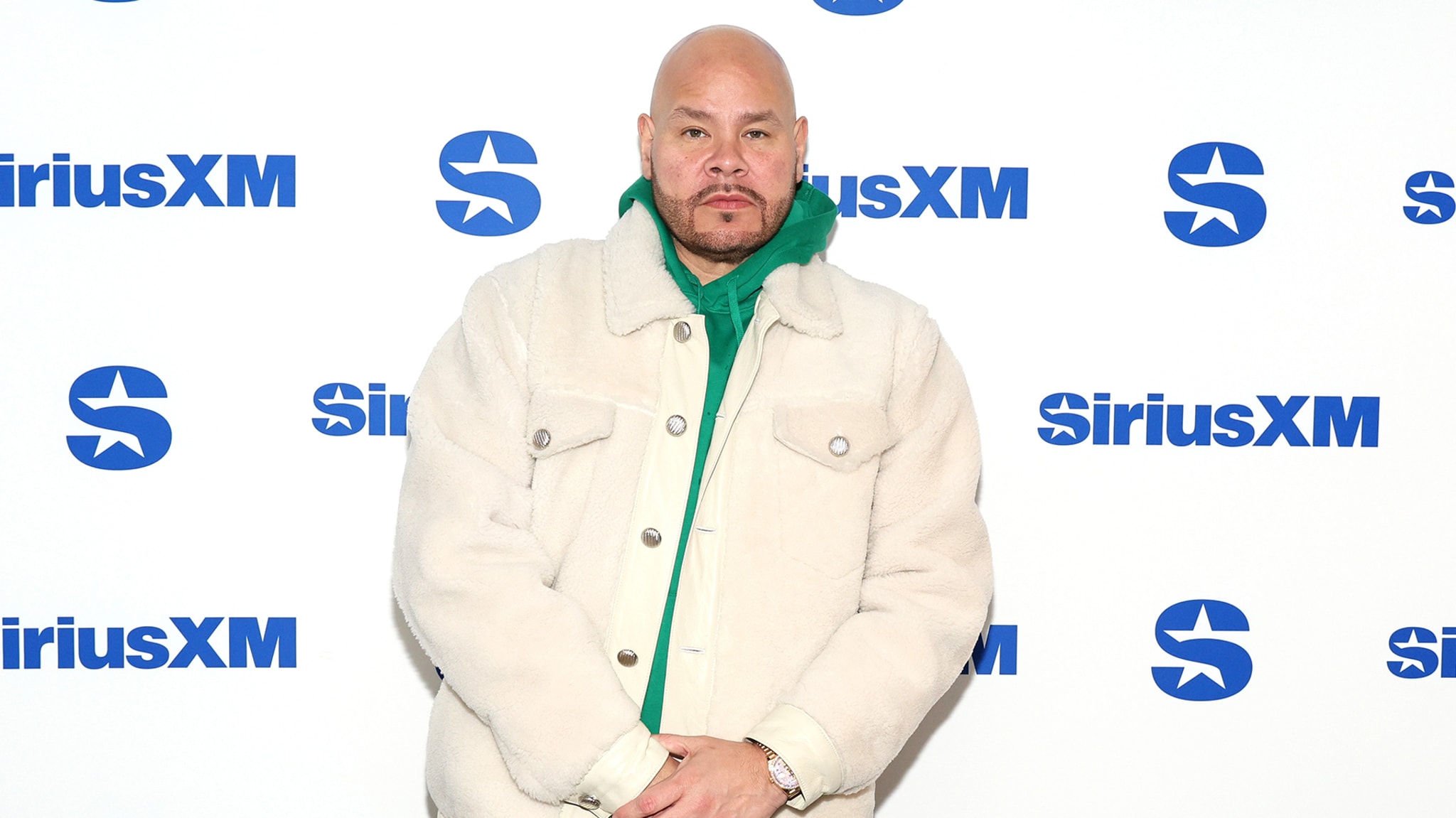 Fat Joe at a SiriusXM event, wearing a white coat over a green hoodie, standing against a branded backdrop.