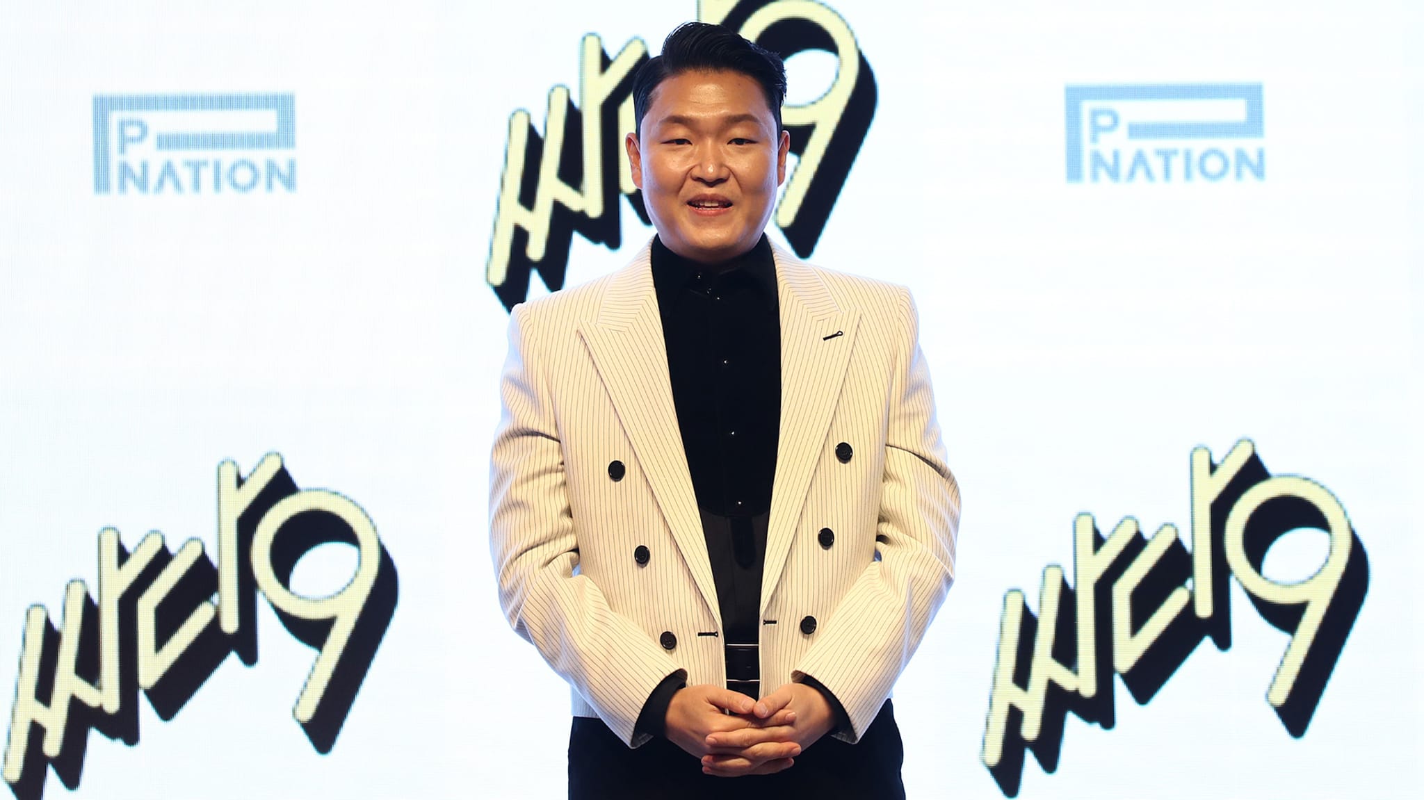 Psy, wearing a striped suit, stands in front of a backdrop with "PNATION" and "9" logos.