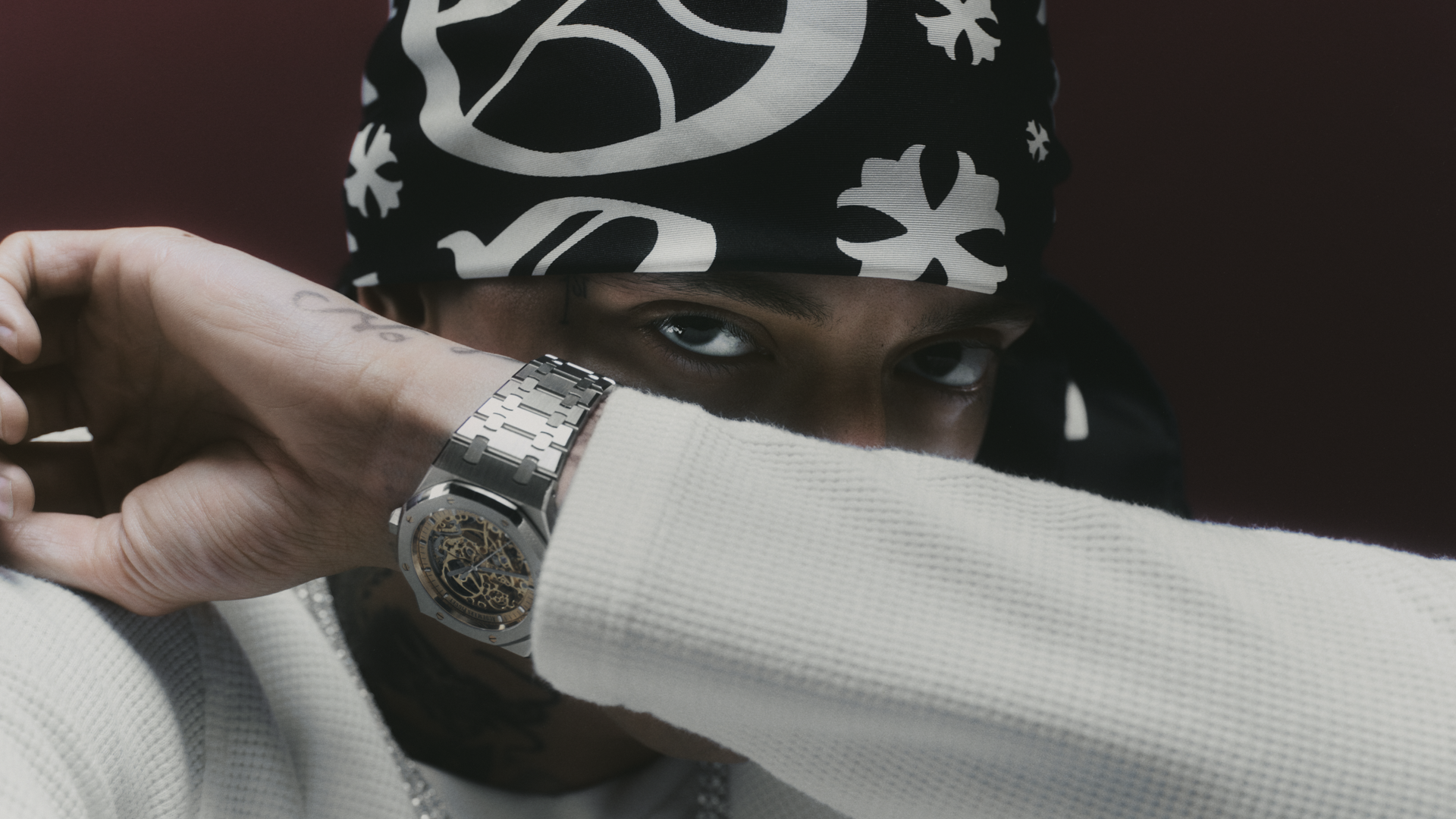 A person wearing a patterned bandana and a silver watch, partially covering their face with their arm, dressed in a white sweater.
