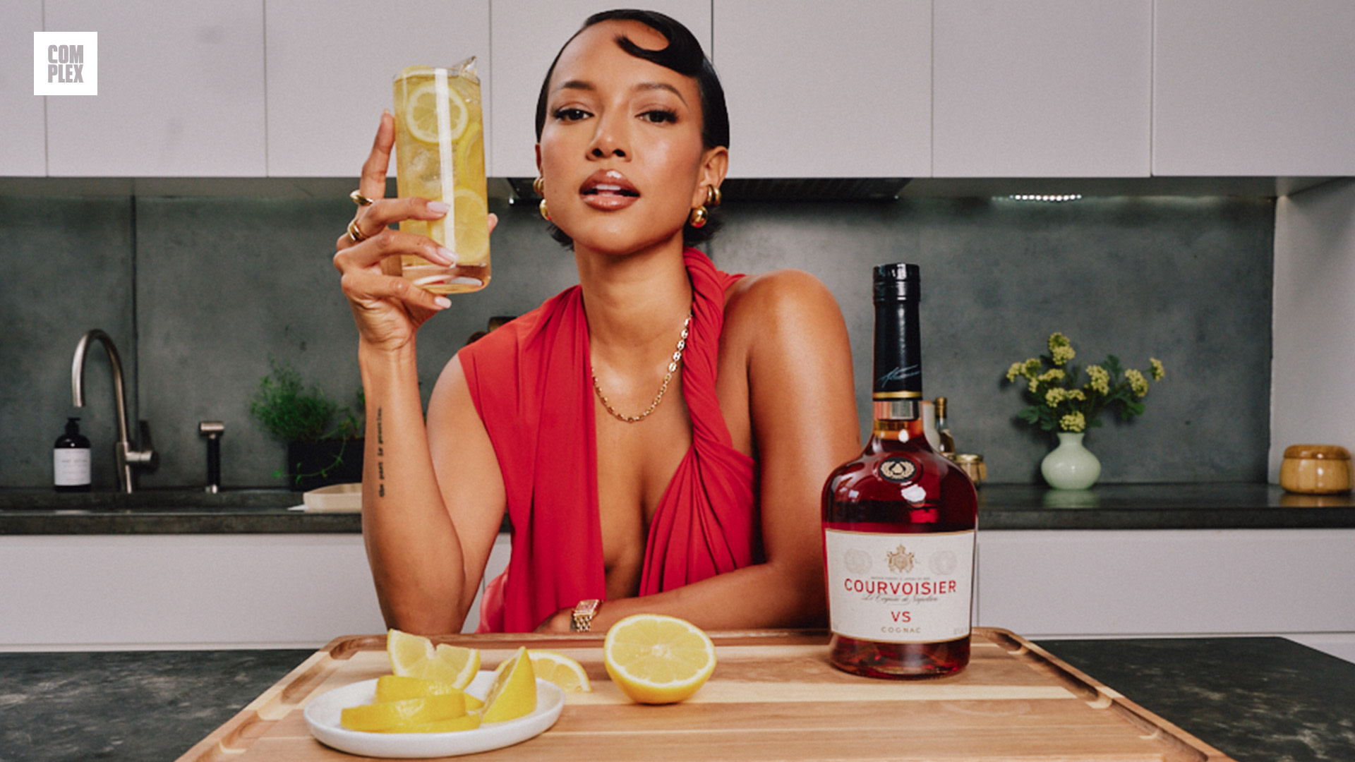 A woman in a red dress holds a lemon drink next to a bottle of Courvoisier on a kitchen counter with lemons.