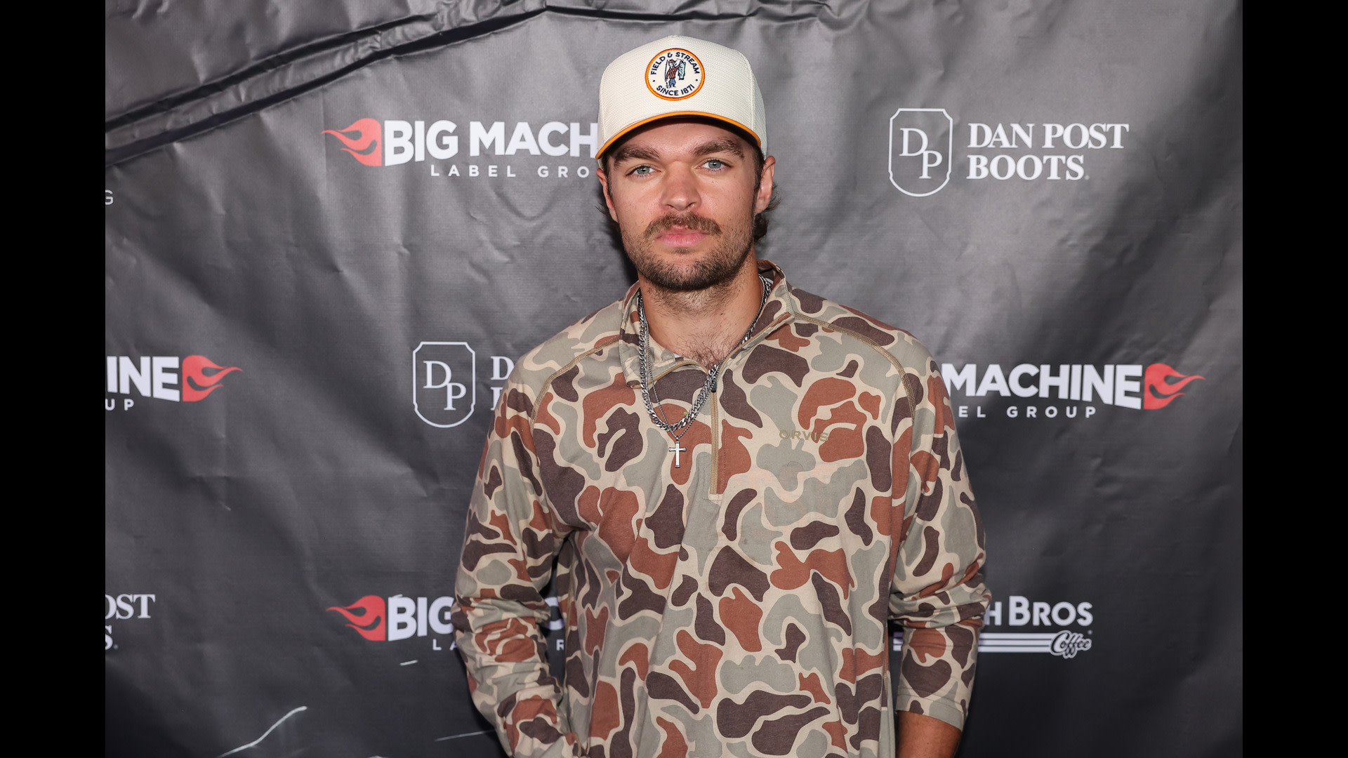 A man in a camo shirt and white cap stands in front of a backdrop with "Big Machine Label Group" and "Dan Post Boots" logos.