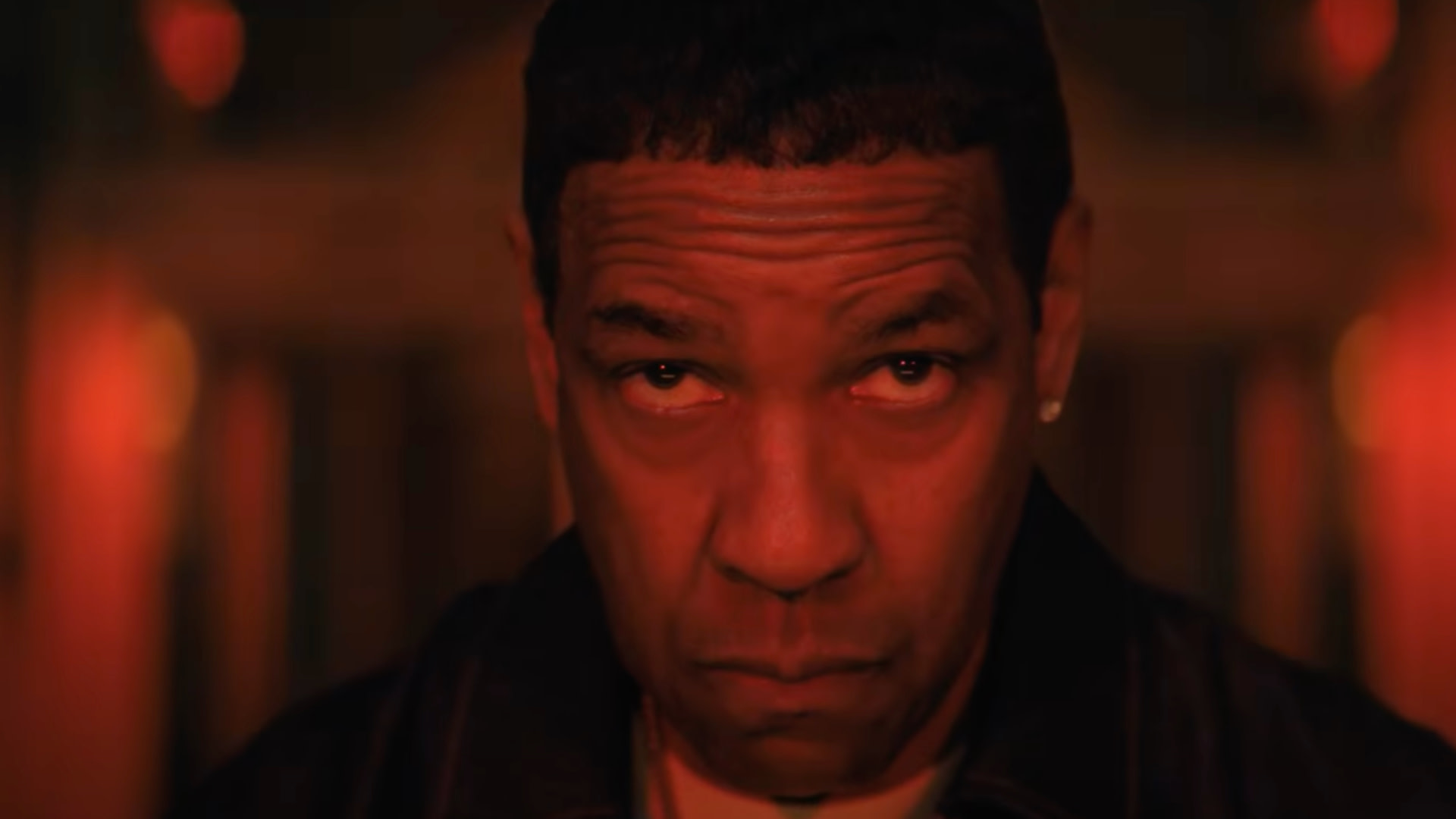 Denzel Washington with an intense expression, lit by a warm, red glow, wearing a dark jacket.