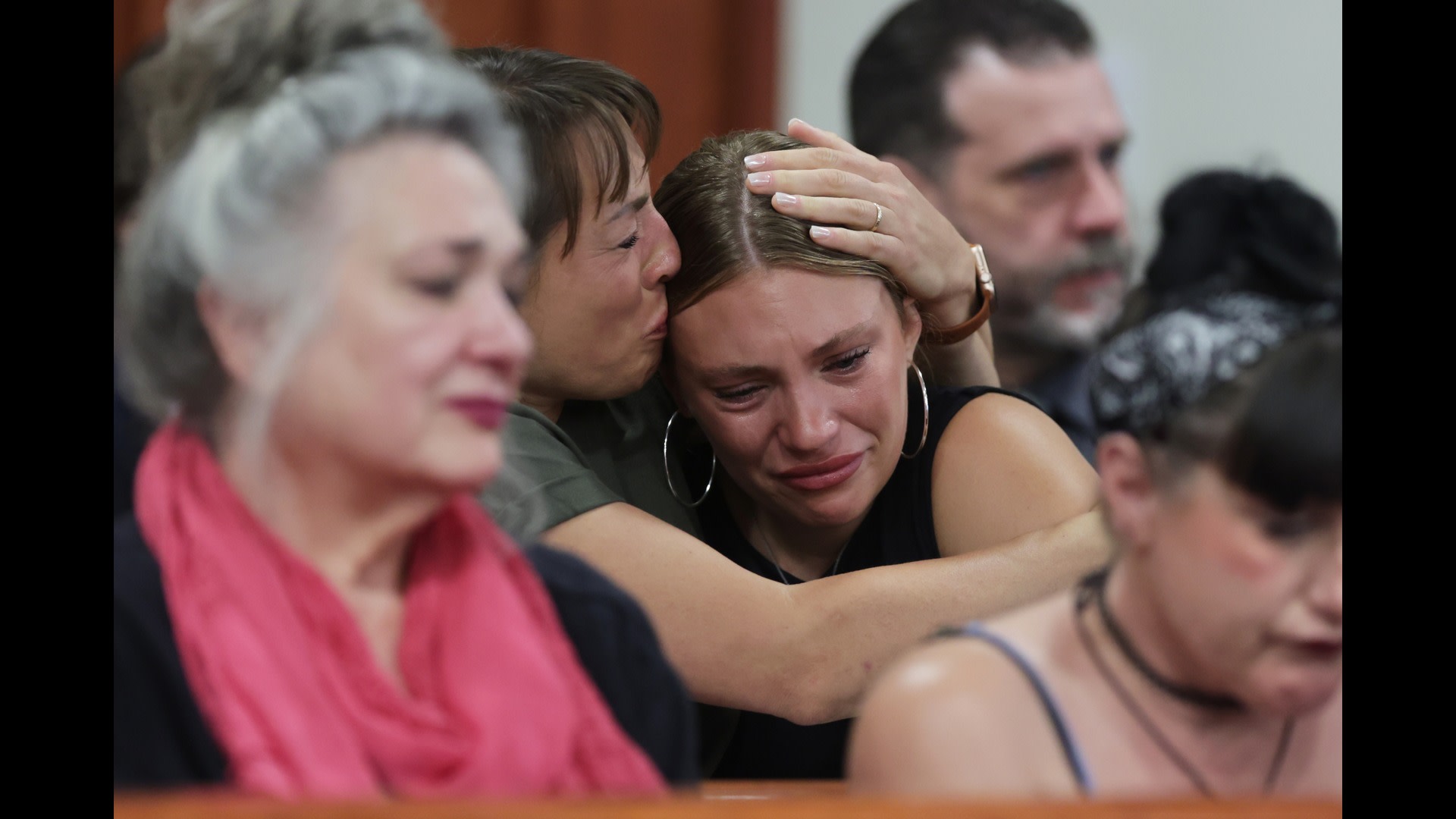 A group of people in a courtroom, some visibly emotional. A woman comforts another who is crying, while others look on somberly.