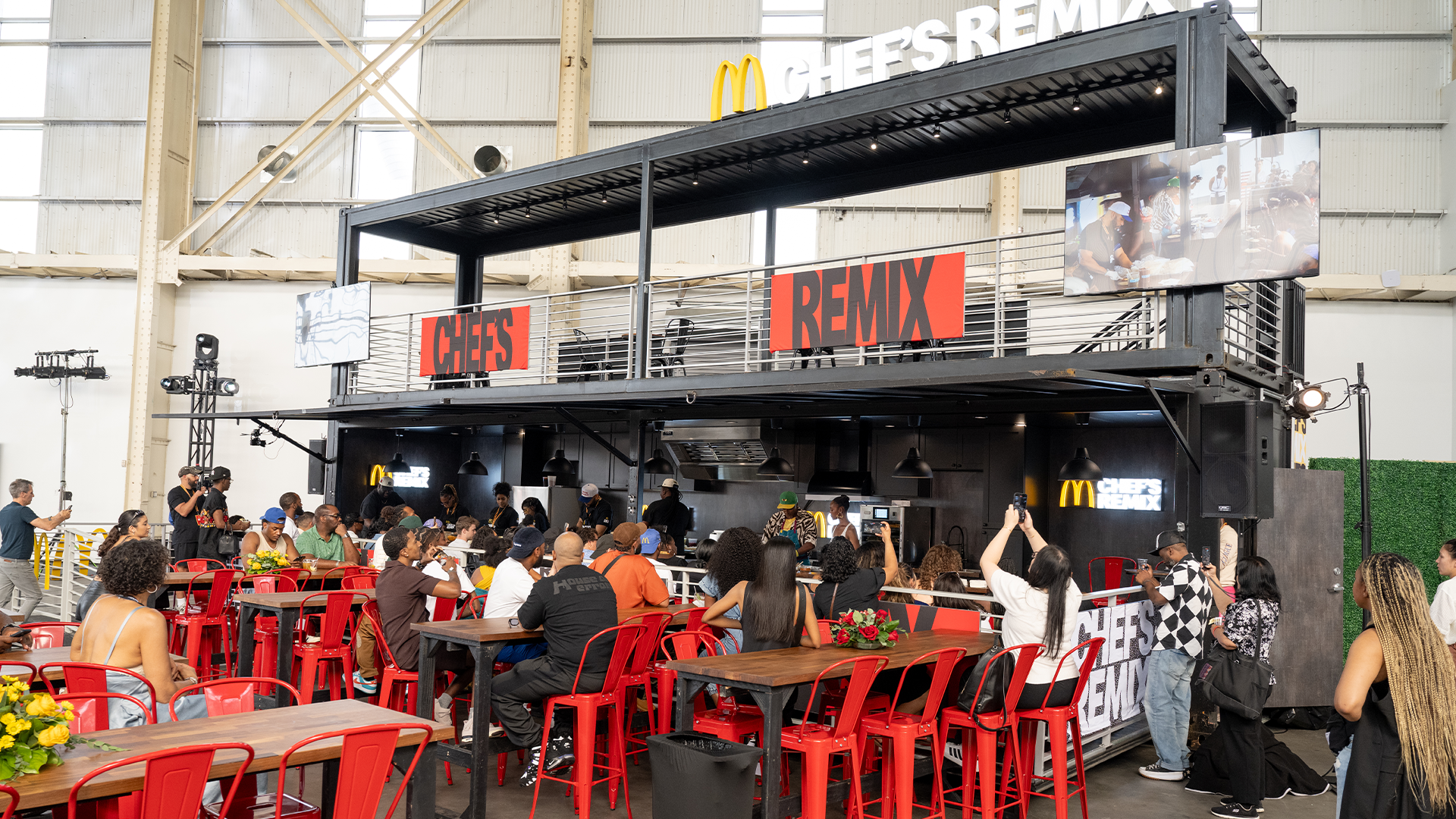 People sitting at tables with red chairs in a McDonald s-themed event space labeled "Chef's Remix," featuring a modern, industrial design.