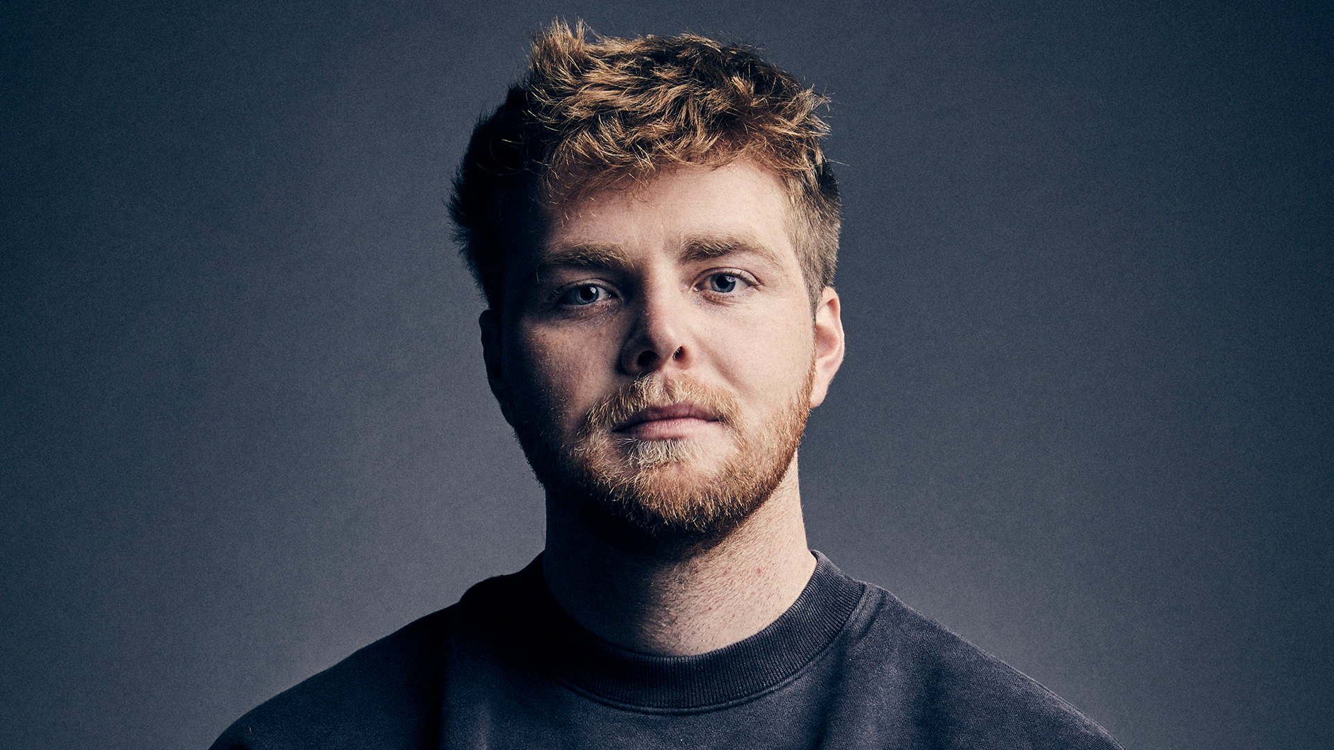 A man with short, tousled hair and a beard, wearing a dark sweatshirt, looks directly at the camera against a plain background.