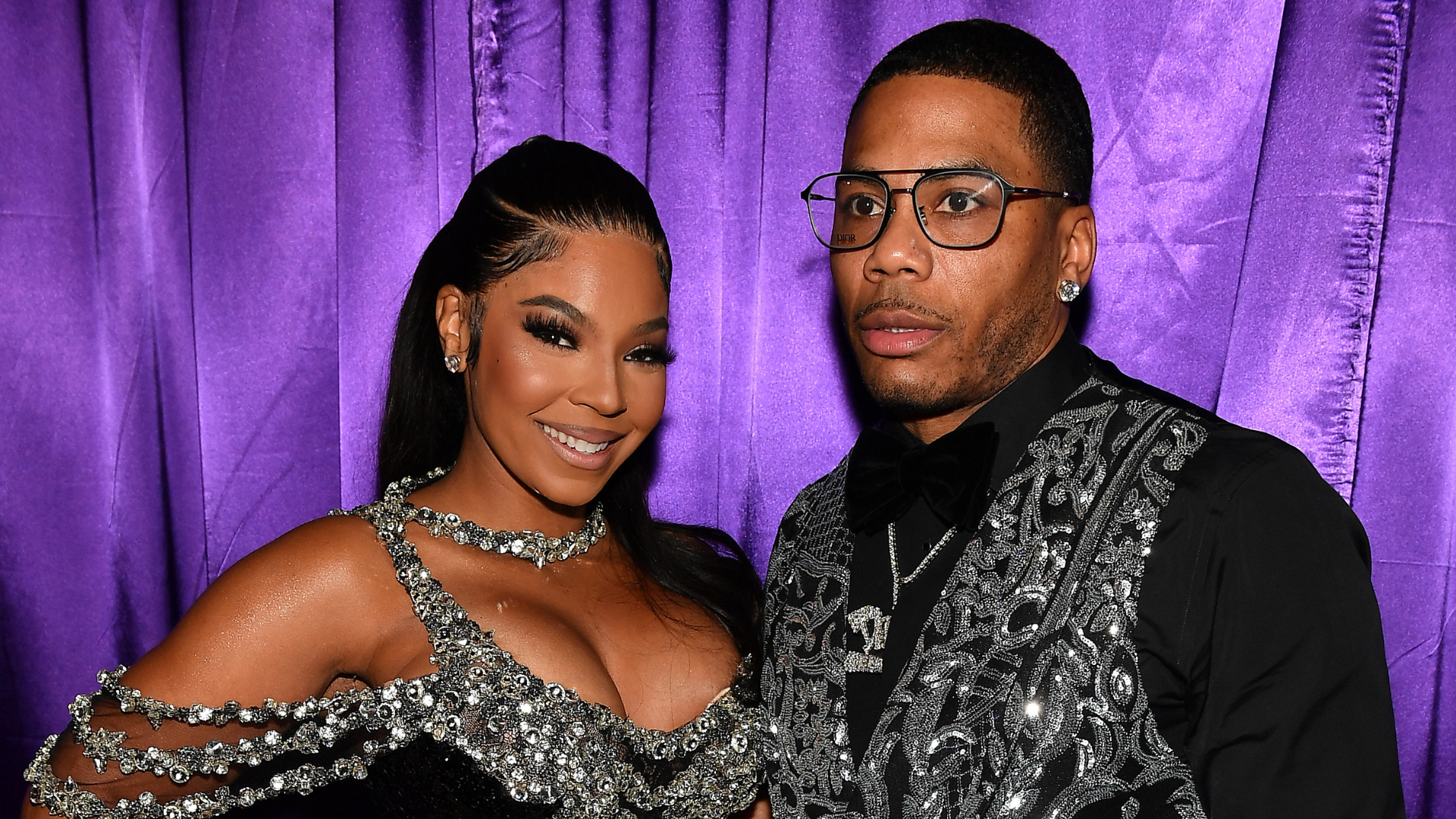 Ashanti and Nelly pose together in glamorous outfits against a purple curtain backdrop.