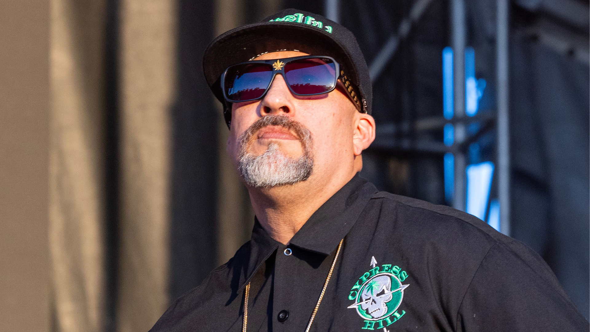 A man wearing sunglasses, a black cap, and a black shirt with "Cypress Hill" logo, standing outdoors at an event.