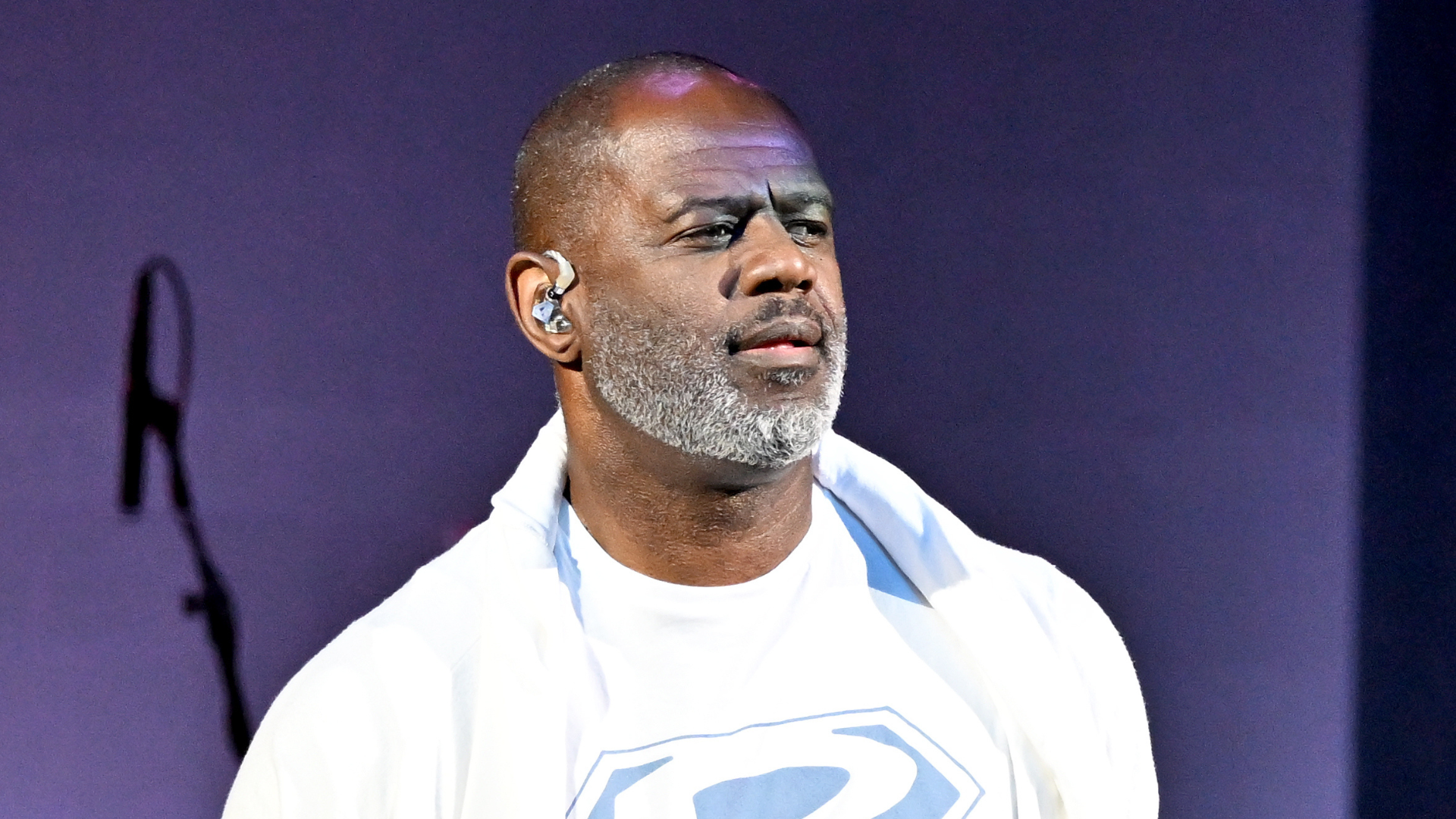 A man with a beard and earpiece stands on stage, wearing a white shirt with a design. The background is purple.