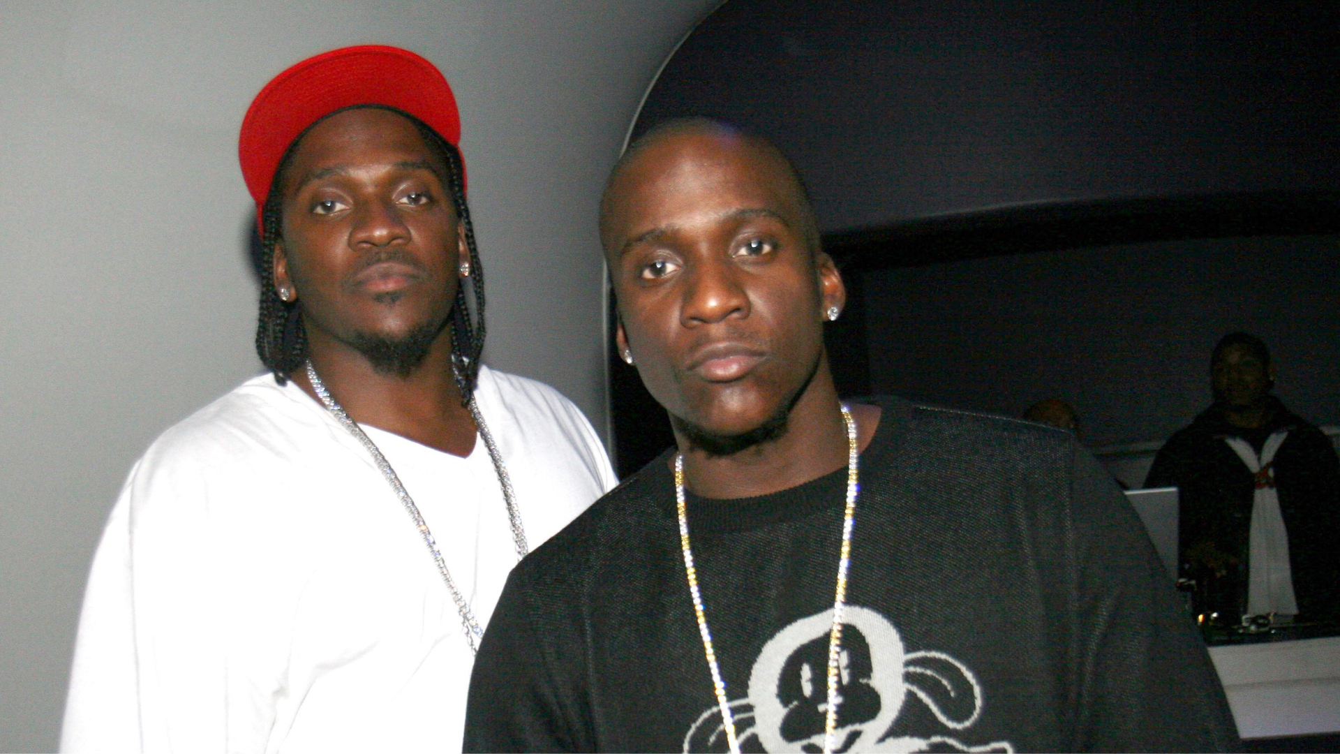 Two men in casual clothing, one wearing a red cap and white shirt, the other in a black shirt with a graphic design.