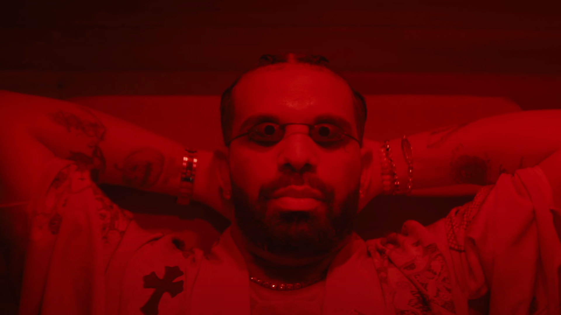 Drake with tattoos and glasses is bathed in red light, wearing a patterned shirt and jewelry, with arms resting behind their head.