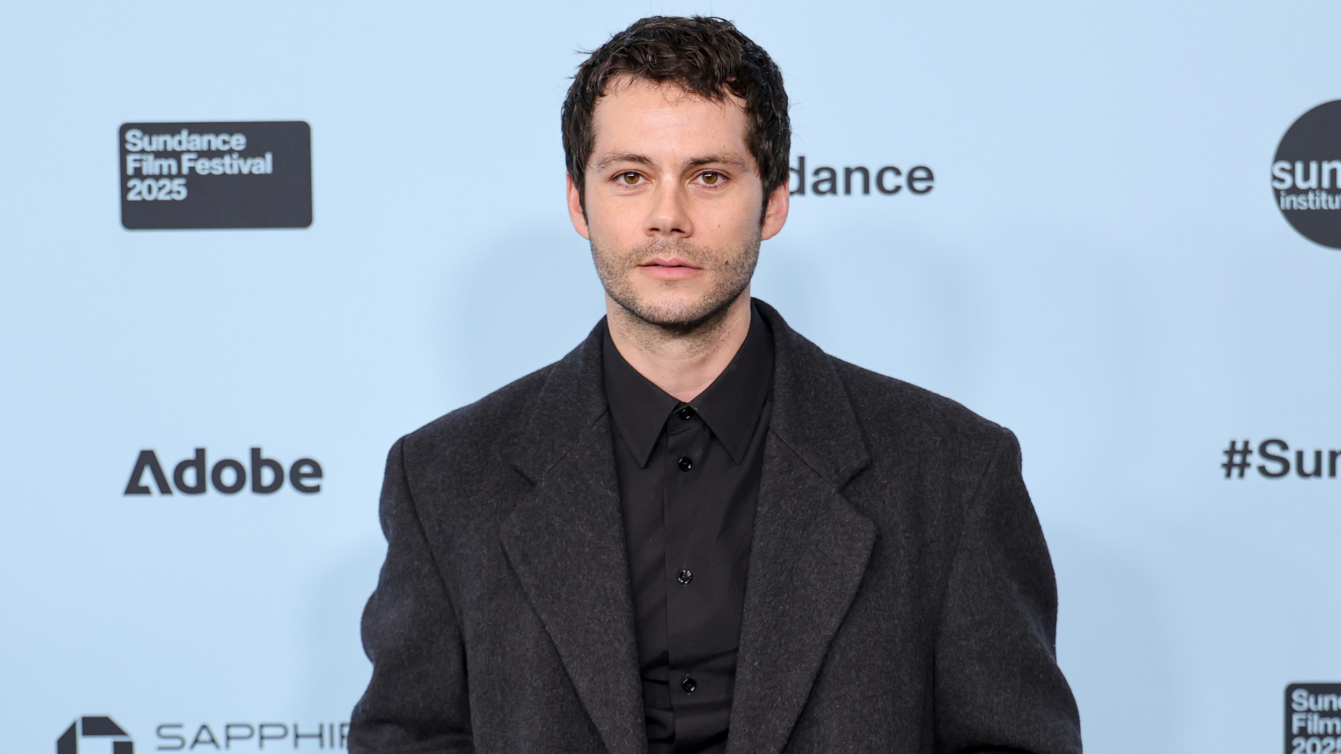 Dylan O'Brien at the Sundance Film Festival 2025, wearing a dark coat and shirt, standing against a light blue backdrop.