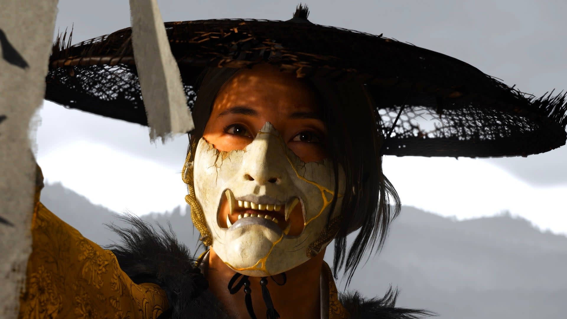 A person wearing a cracked, white mask with fangs and a wide-brimmed hat, set against a mountainous background.