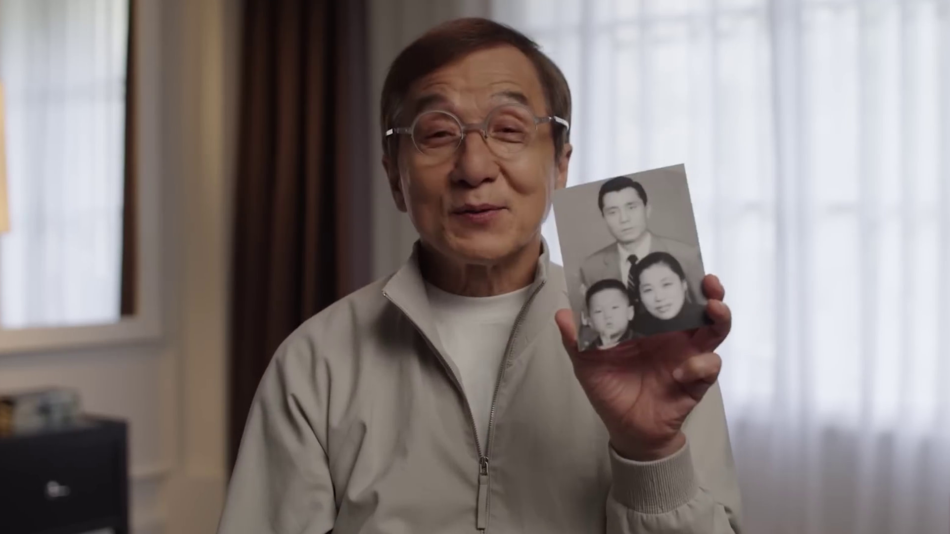 Jackie Chan wearing glasses holds a black-and-white family photo, smiling. The room is softly lit with curtains in the background.