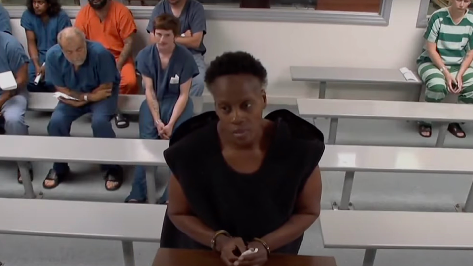 A person stands at a courtroom podium wearing a black outfit. Several people sit on benches in the background, some in orange or striped uniforms.