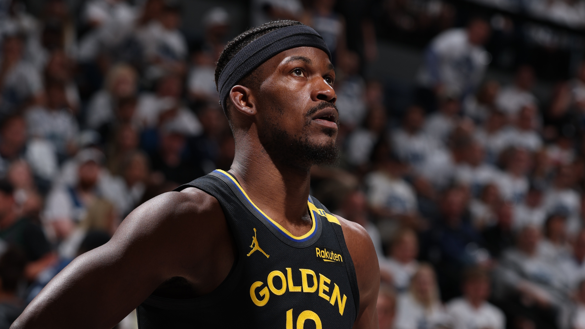 Jimmy Butler in a black "Golden" jersey with No. 10, wearing a headband, on a court with a blurred crowd in the background.