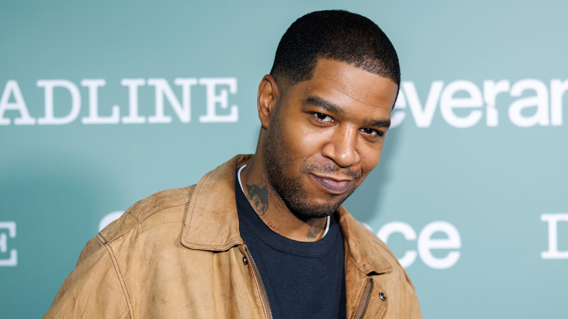 A man in a tan jacket and black shirt stands against a green backdrop with white text.