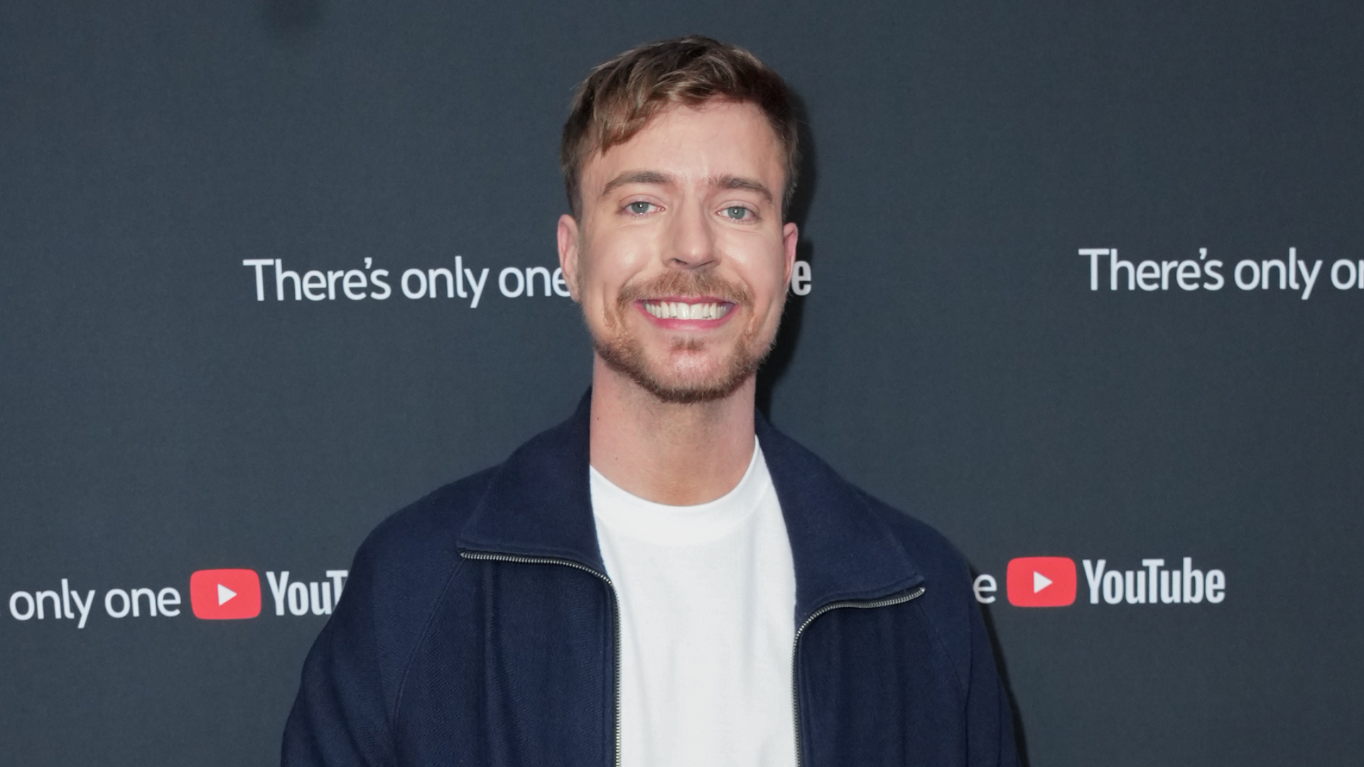 A man with short hair and a beard smiles at a YouTube event, standing against a backdrop with the YouTube logo.