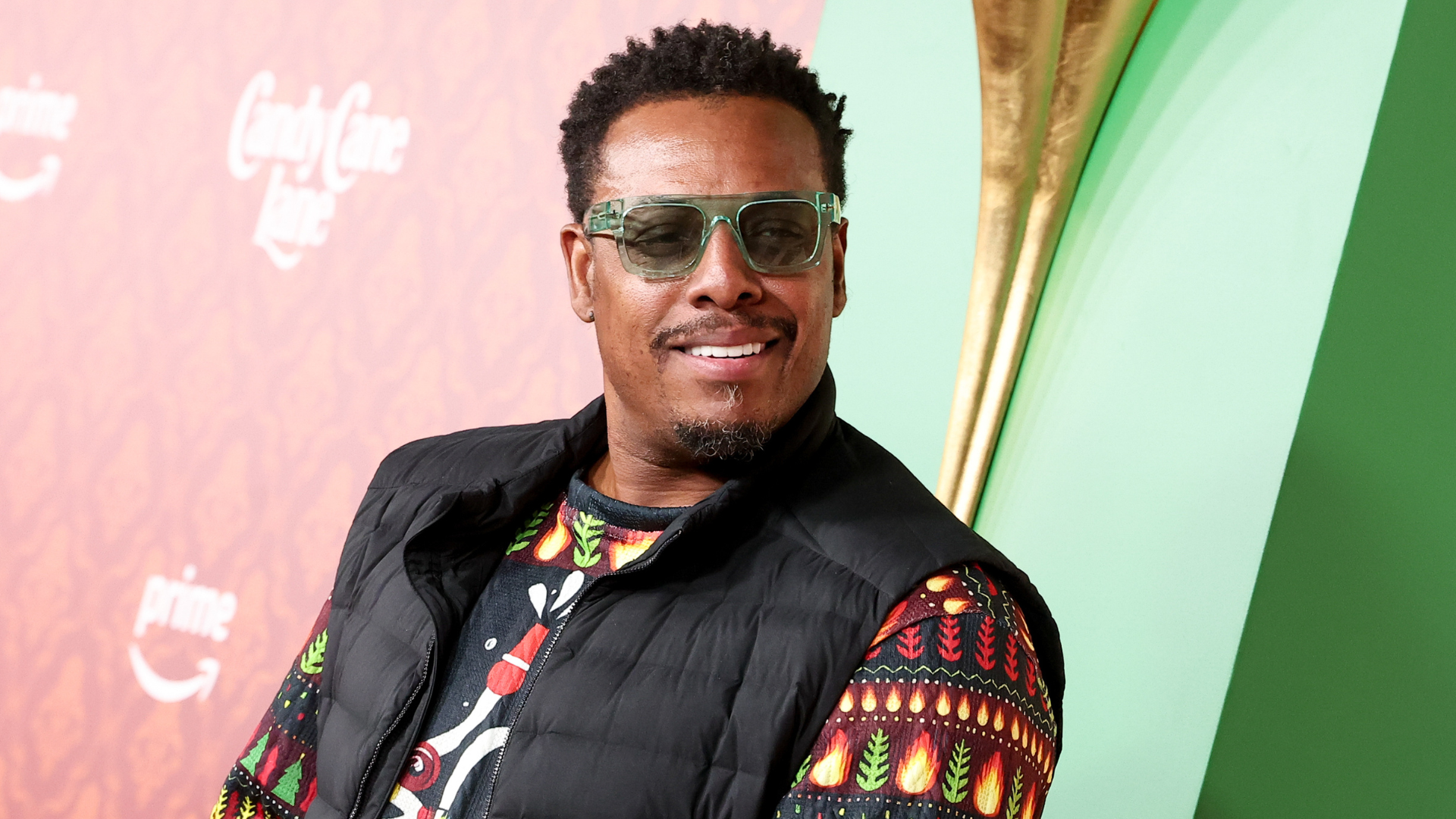 A man wearing sunglasses and a colorful shirt with a black vest, smiling at an event with a red and green background.