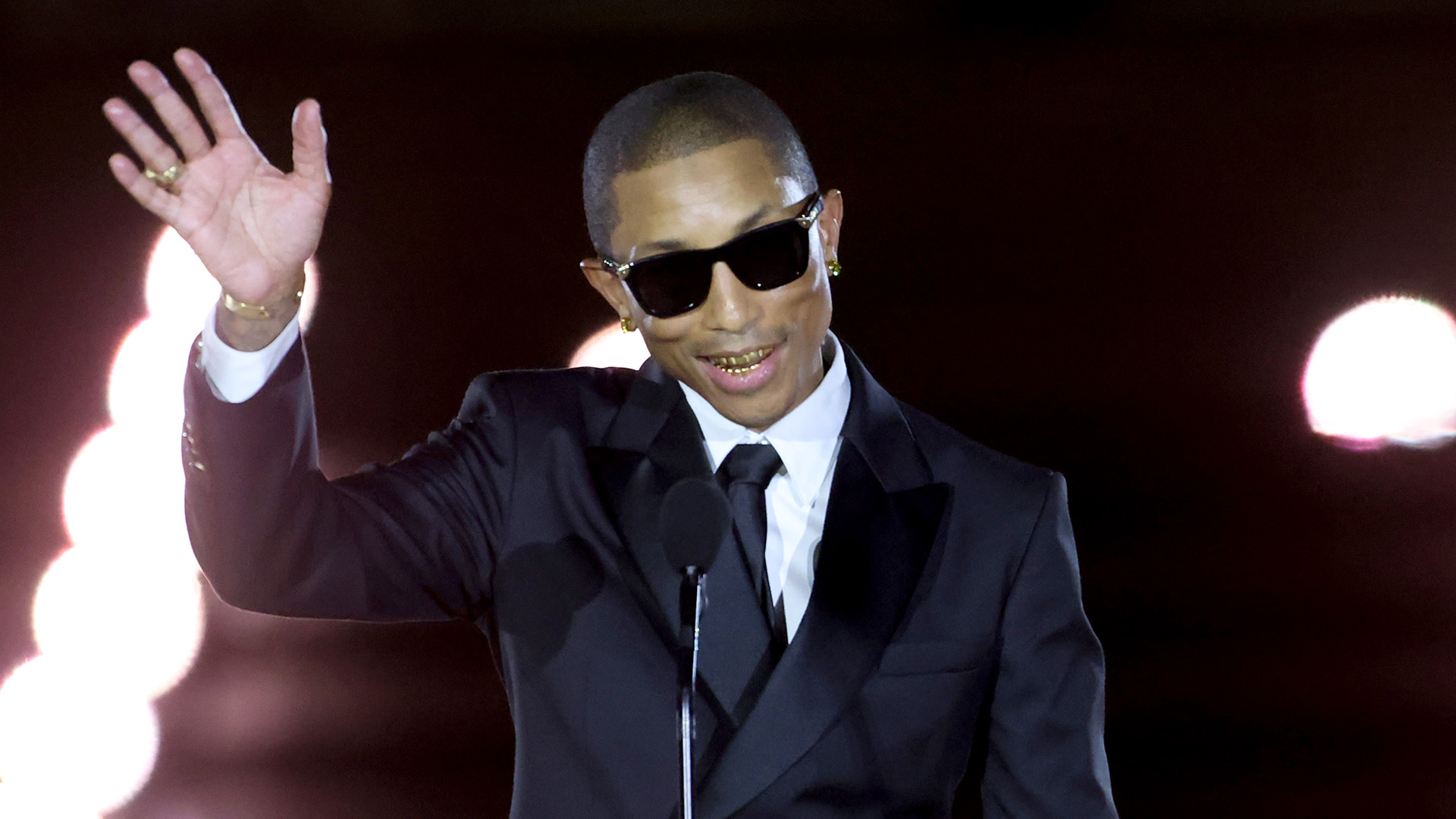 Pharrell Williams, in a suit and sunglasses, waves while speaking at a microphone.