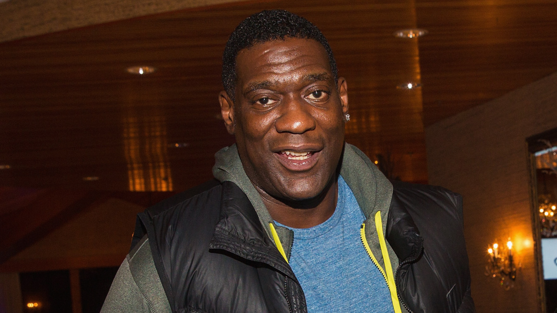 A man wearing a blue shirt and black vest smiles indoors, with warm lighting in the background.
