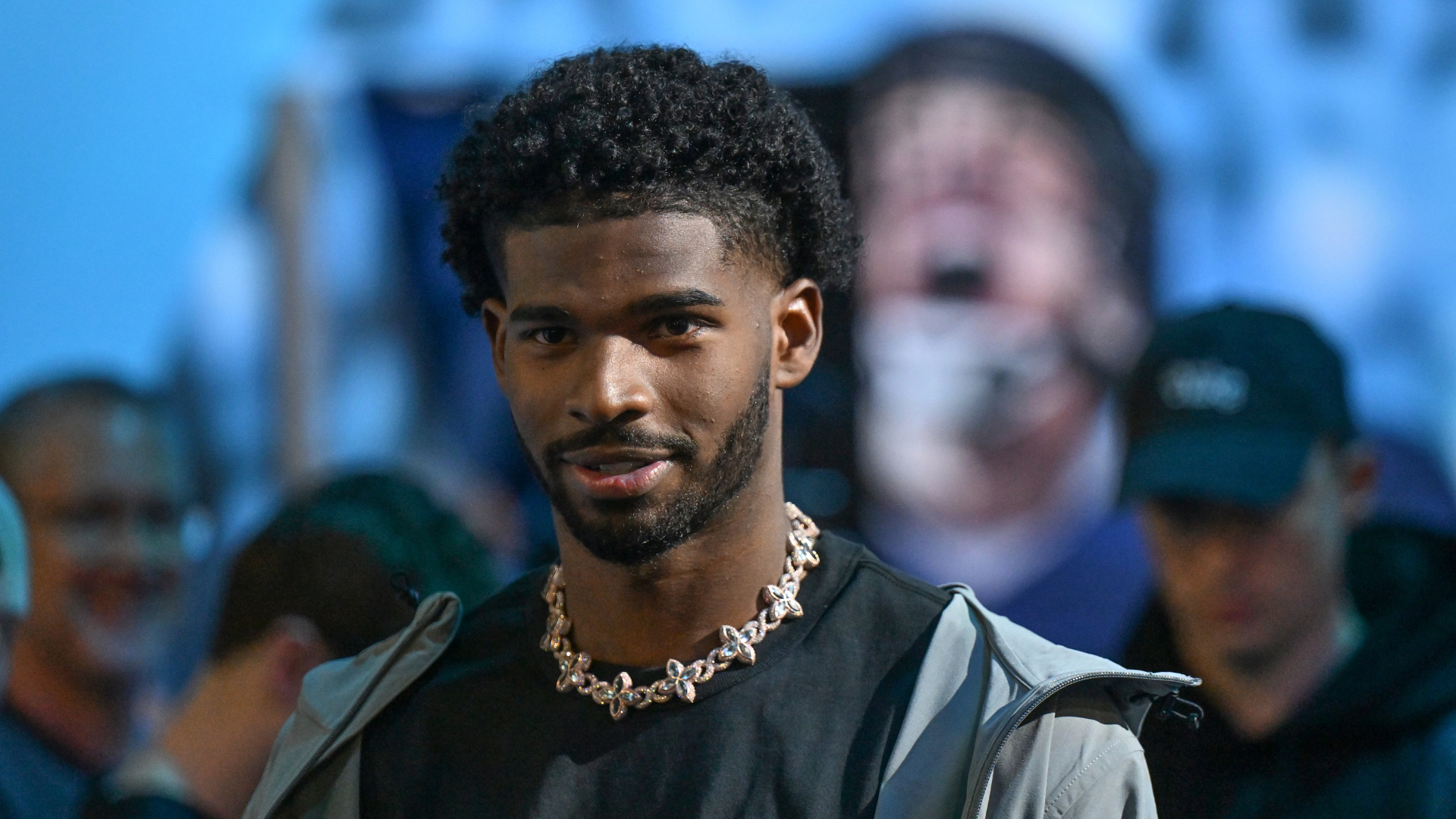 A man with short curly hair and a beard, wearing a black shirt, silver chain, and a light jacket, stands in a crowd.