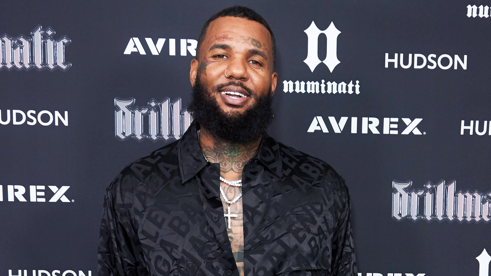 The image shows a man with tattoos and a beard, wearing a patterned black shirt, standing in front of a branded backdrop.