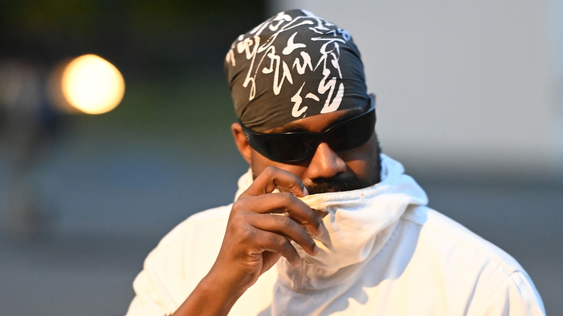A person wearing sunglasses and a patterned bandana, covering their mouth with a white garment.