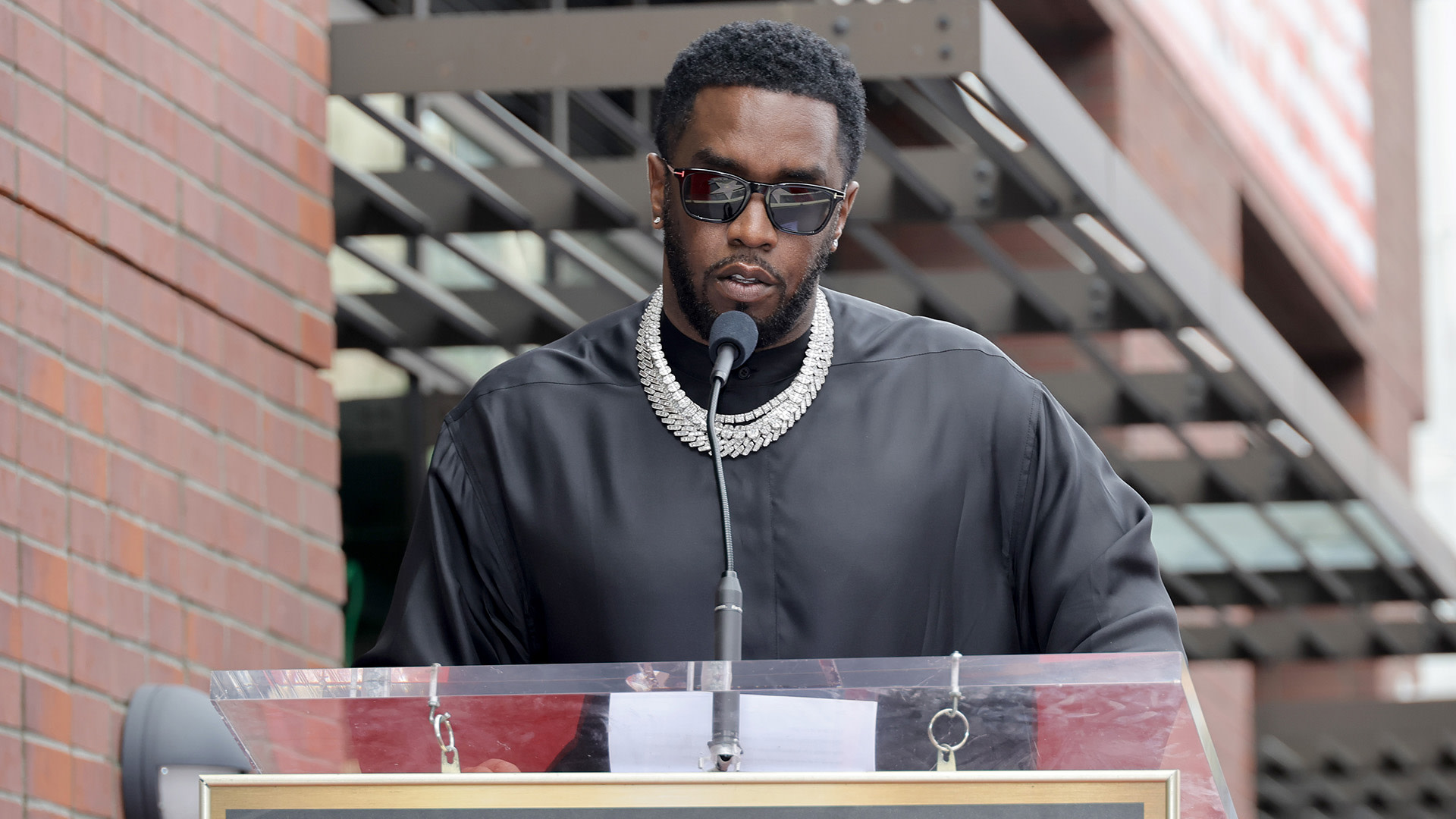 Sean "Diddy" Combs speaking at a podium, wearing sunglasses and a black outfit with a diamond necklace.