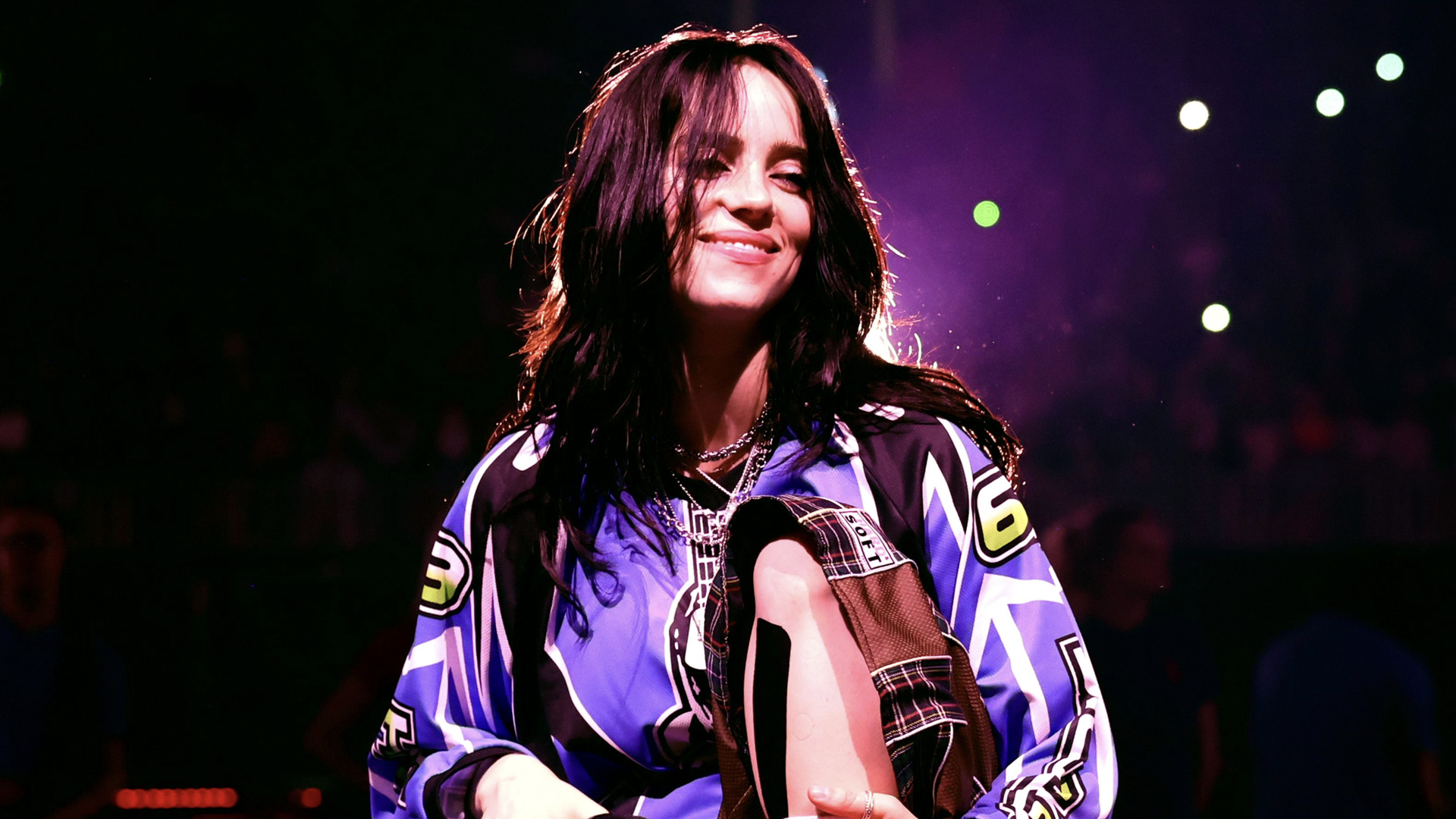 Billie Eilish performing on stage, wearing a colorful jersey, smiling with a spotlight behind her.
