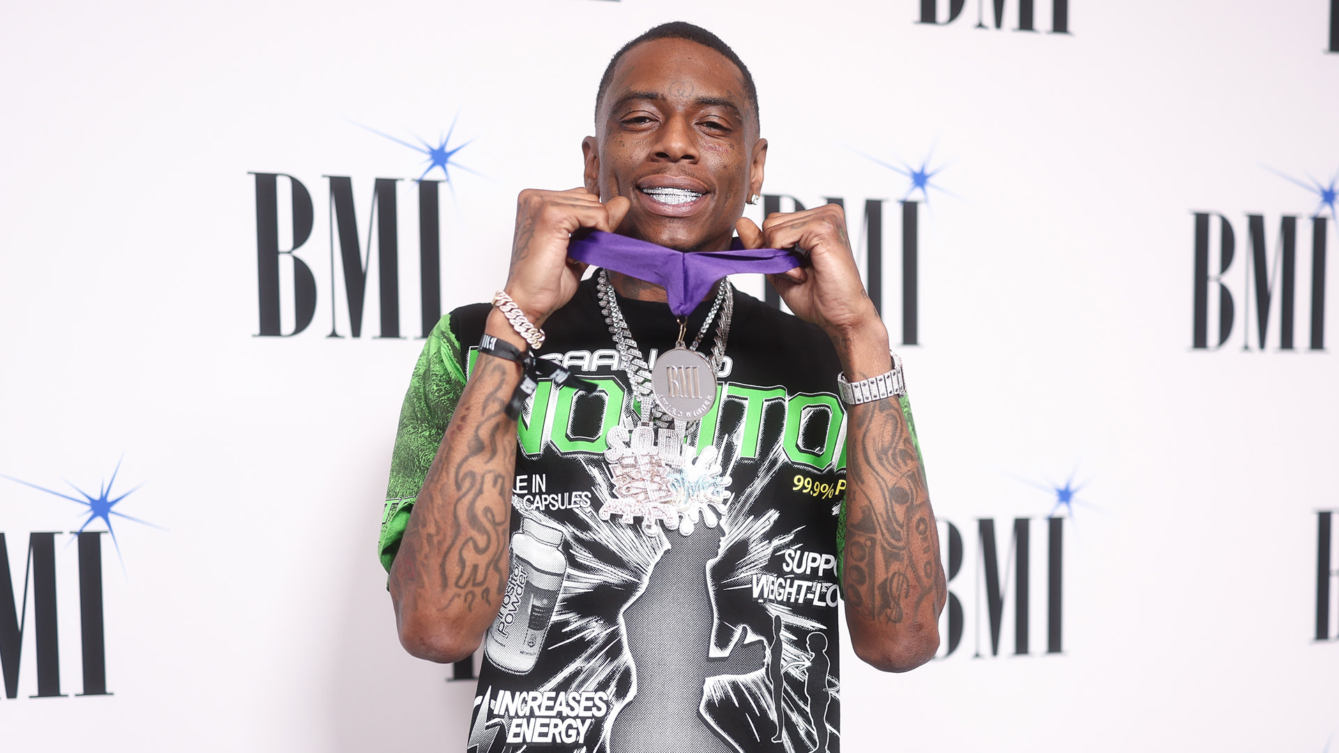 Rapper Soulja Boy at a BMI event, wearing a graphic T-shirt and holding a purple ribbon, with a large chain necklace.