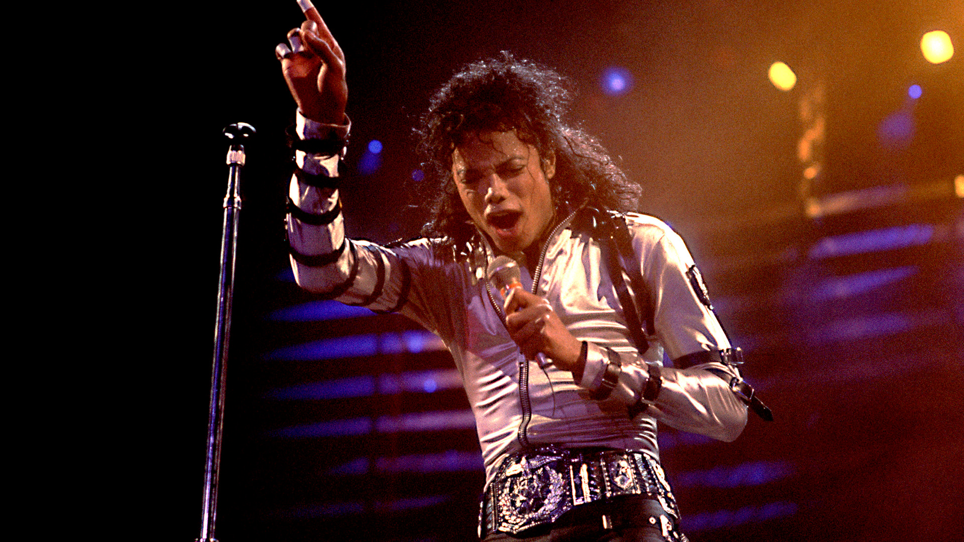 Michael Jackson performing on stage, wearing a white jacket and black belt, with one arm raised and holding a microphone.
