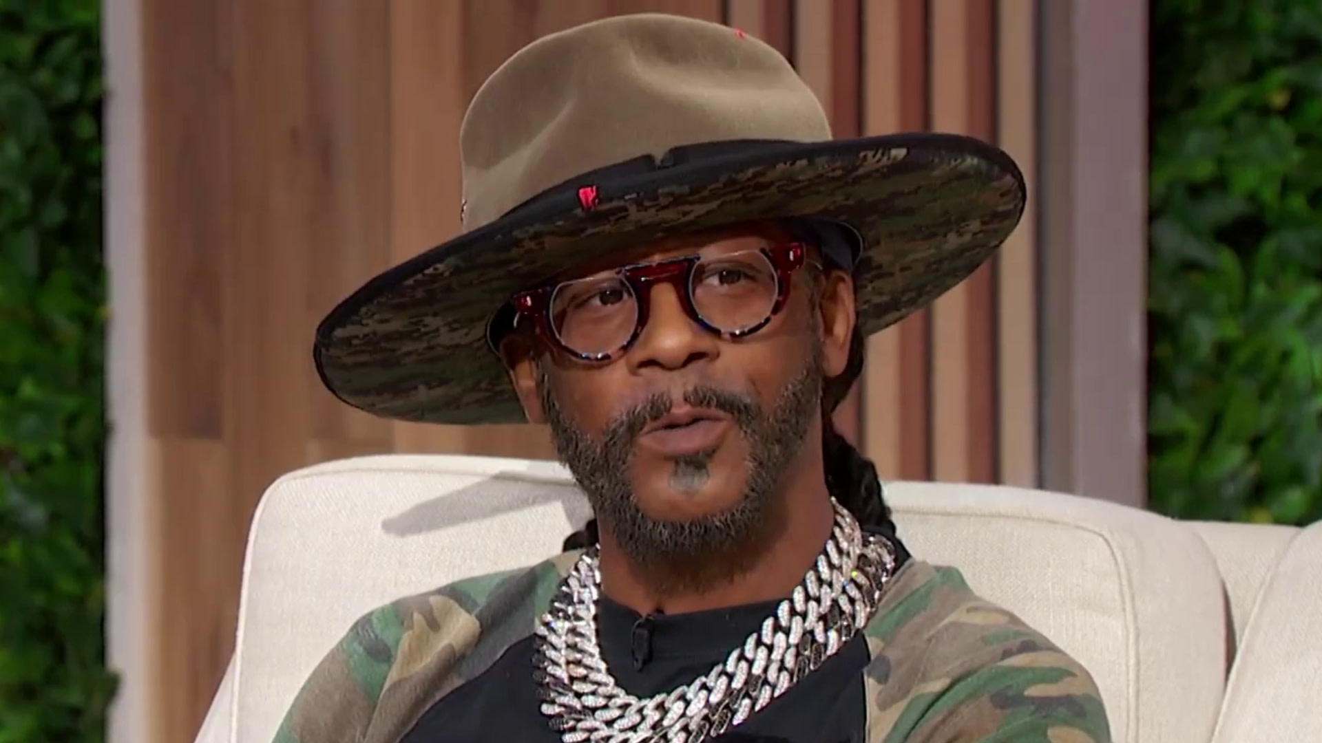 Katt Williams wearing a wide-brimmed hat, glasses, and layered chains, seated on a couch.