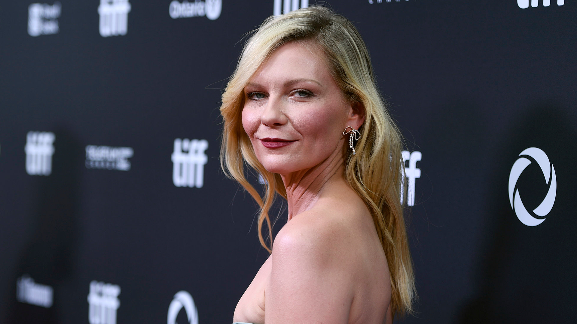 Kirsten Dunst at an event, wearing a strapless dress, with long blonde hair and elegant earrings, standing against a dark background.