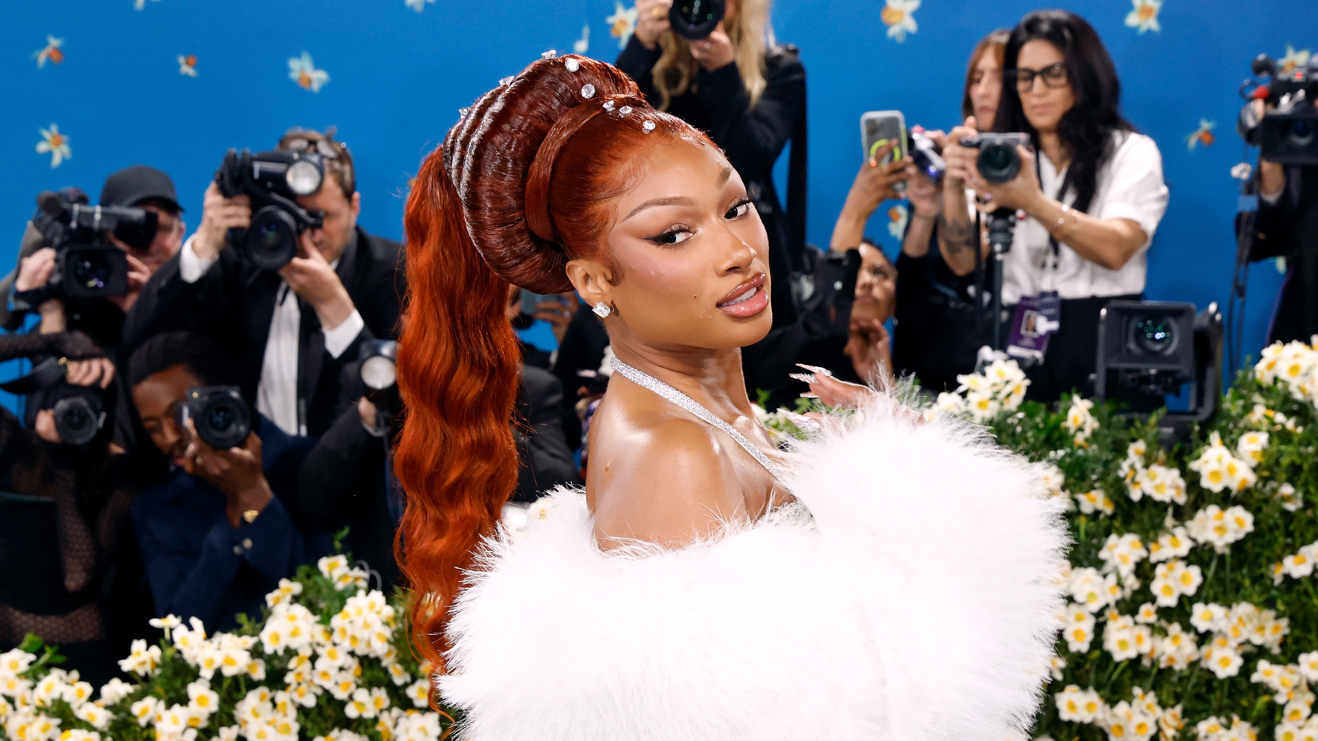 Megan Thee Stallion poses in a glamorous white outfit with a floral background, surrounded by photographers.