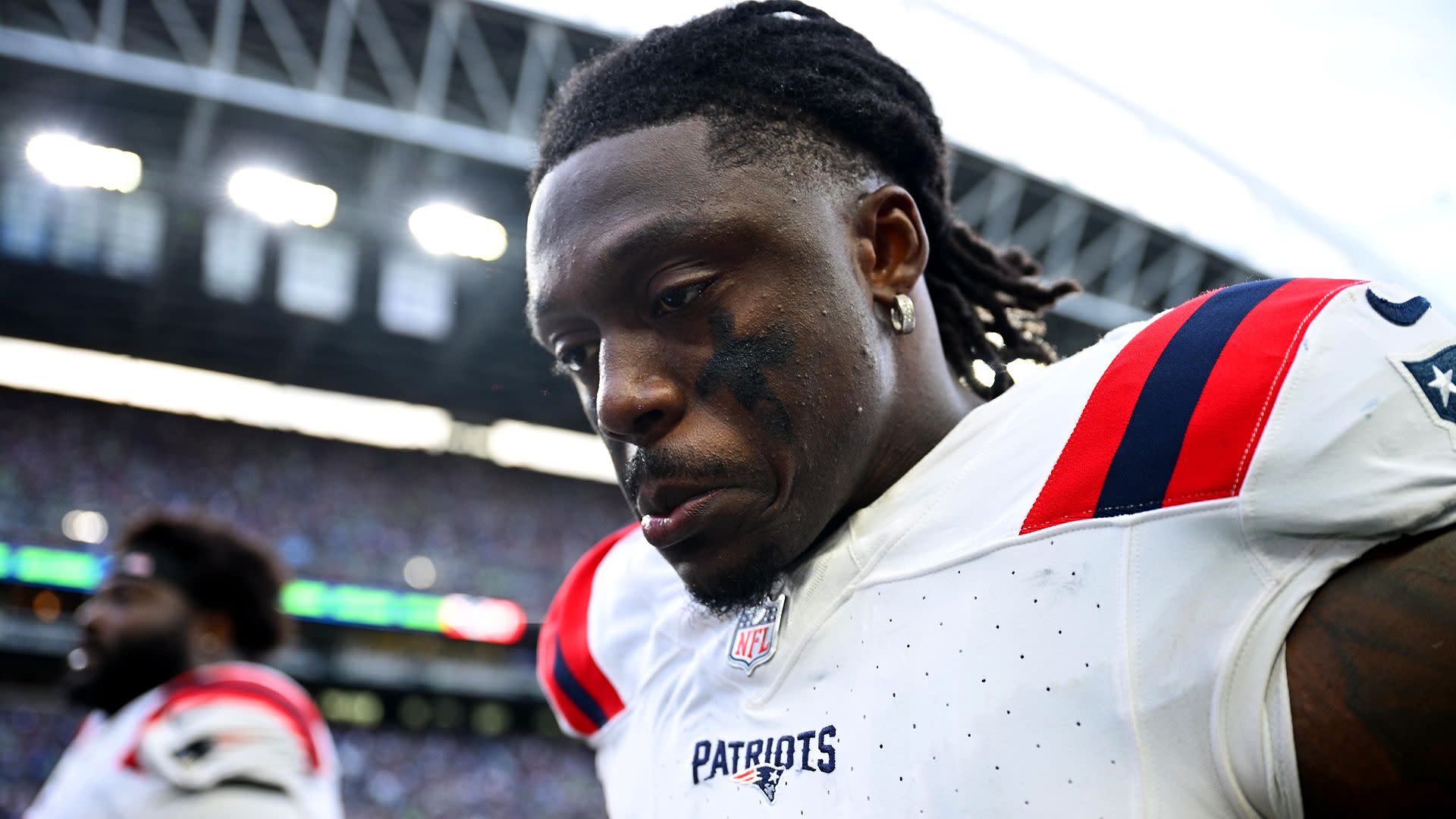 A.J. Brown a New England Patriots jersey on the field, looking down with a focused expression. Stadium and crowd in the background.