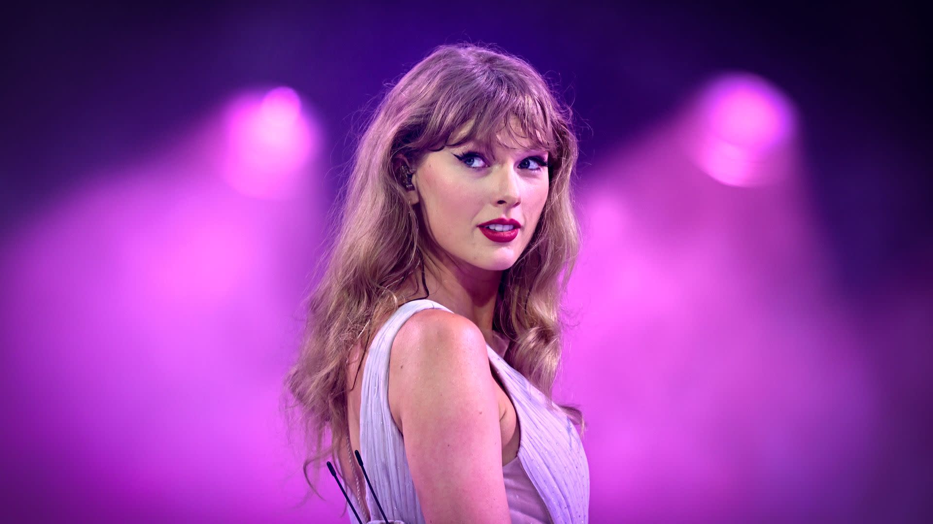 Taylor Swift performing on stage, wearing a light-colored dress, with a purple-lit background.